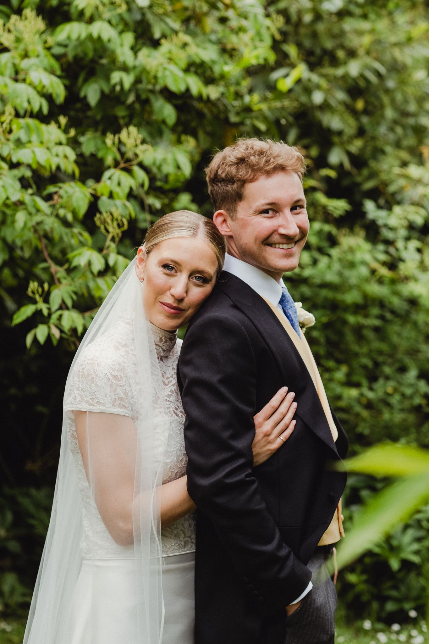 A bride and groom pose together outdoors against lush green foliage during their wedding celebration.