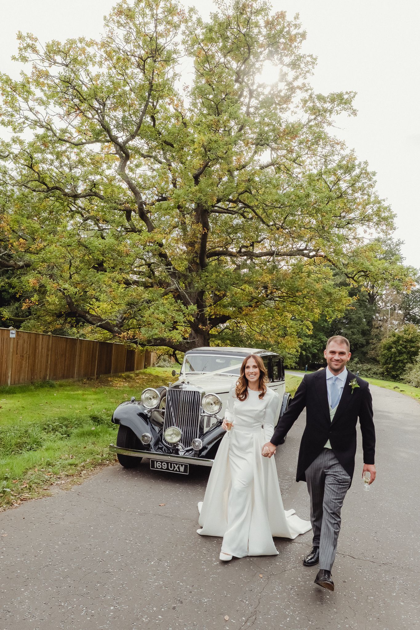 A couple in wedding attire walks down a tree-lined path with a vintage car visible in the background.