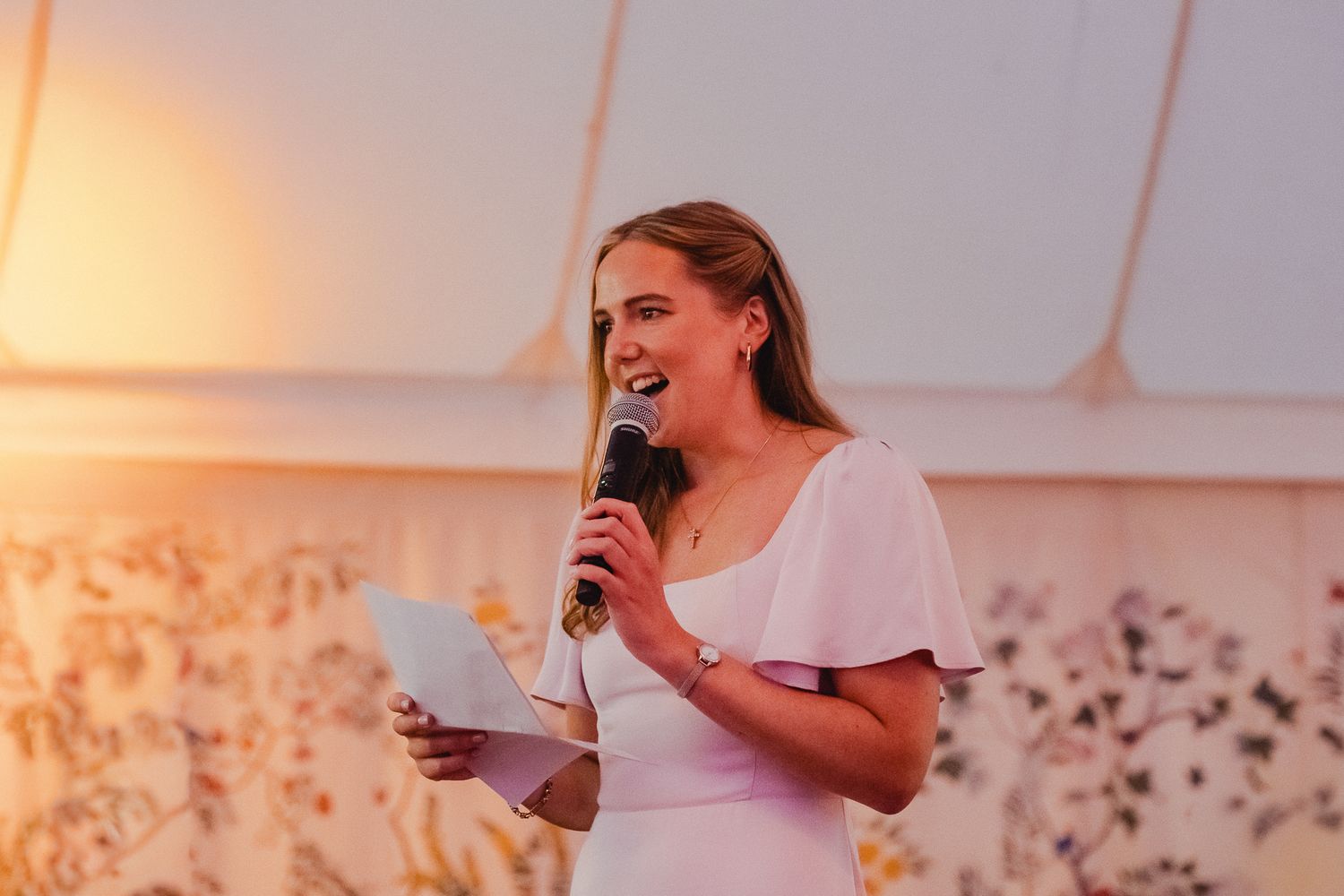A speaker in a white dress holds a microphone and papers while speaking at an event with warm lighting.