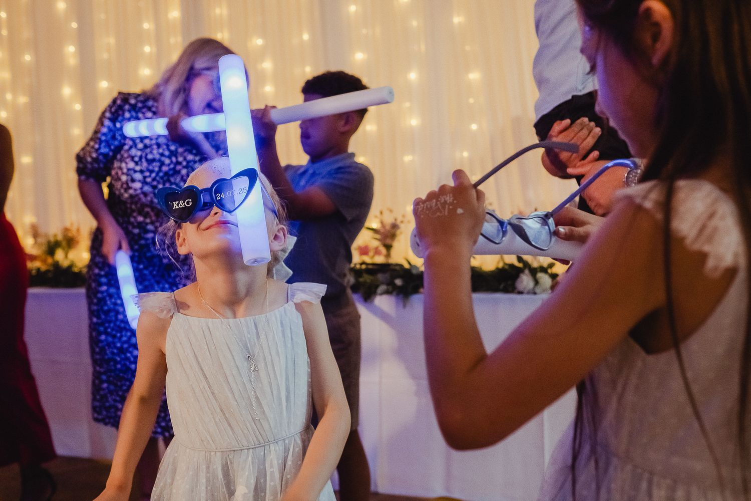 People dancing and celebrating at a wedding reception with glowing string lights in the background.