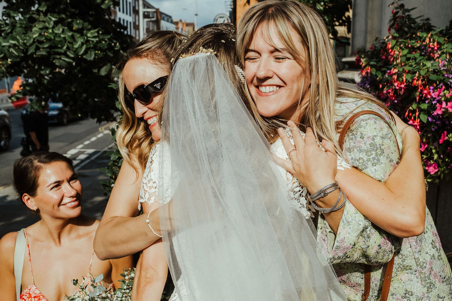 A candid moment at an outdoor wedding celebration with guests and a bride in a flowing veil on a sunny day.