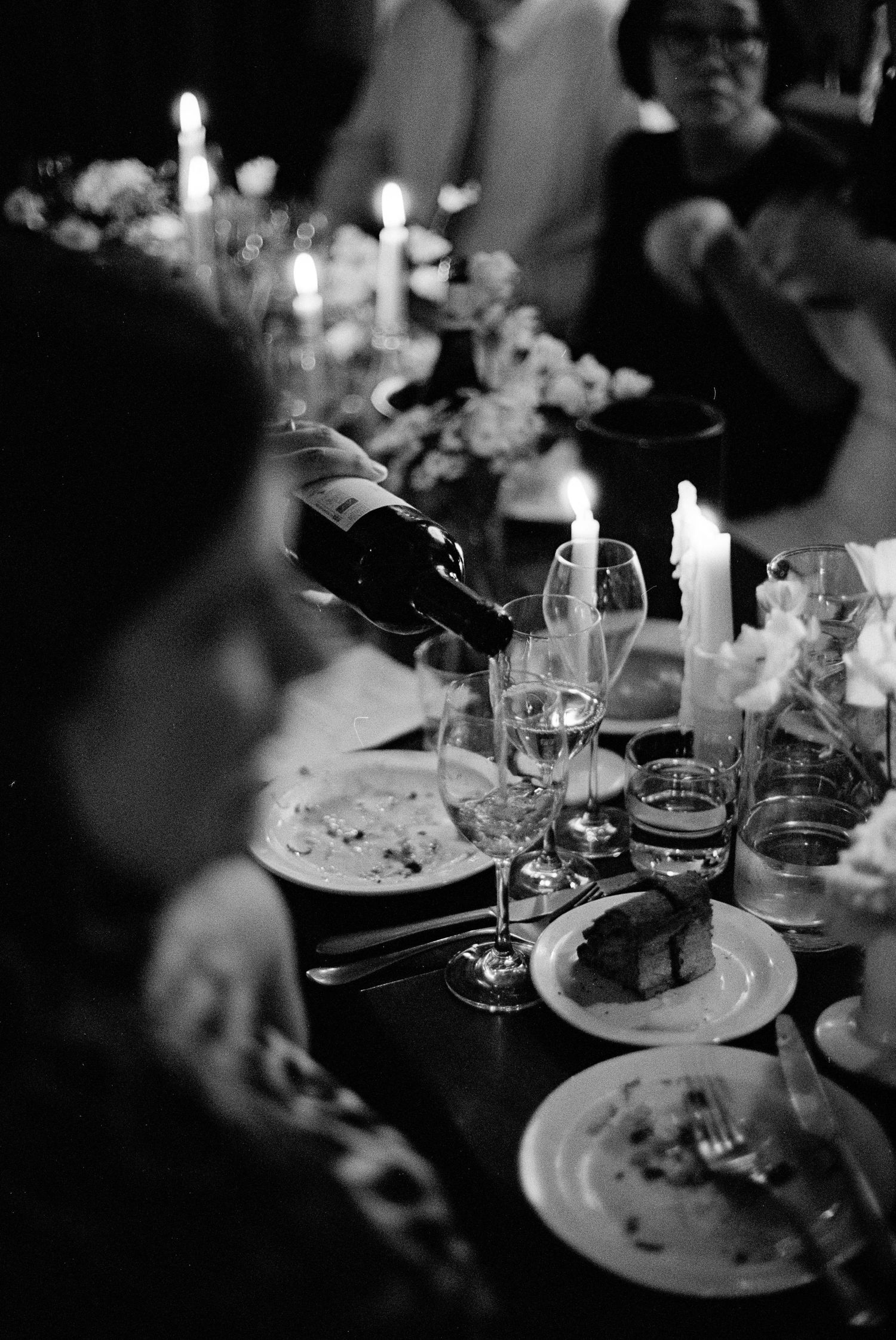 Black and white photo of an elegant candlelit dinner party with wine being poured into glasses on a formal table.