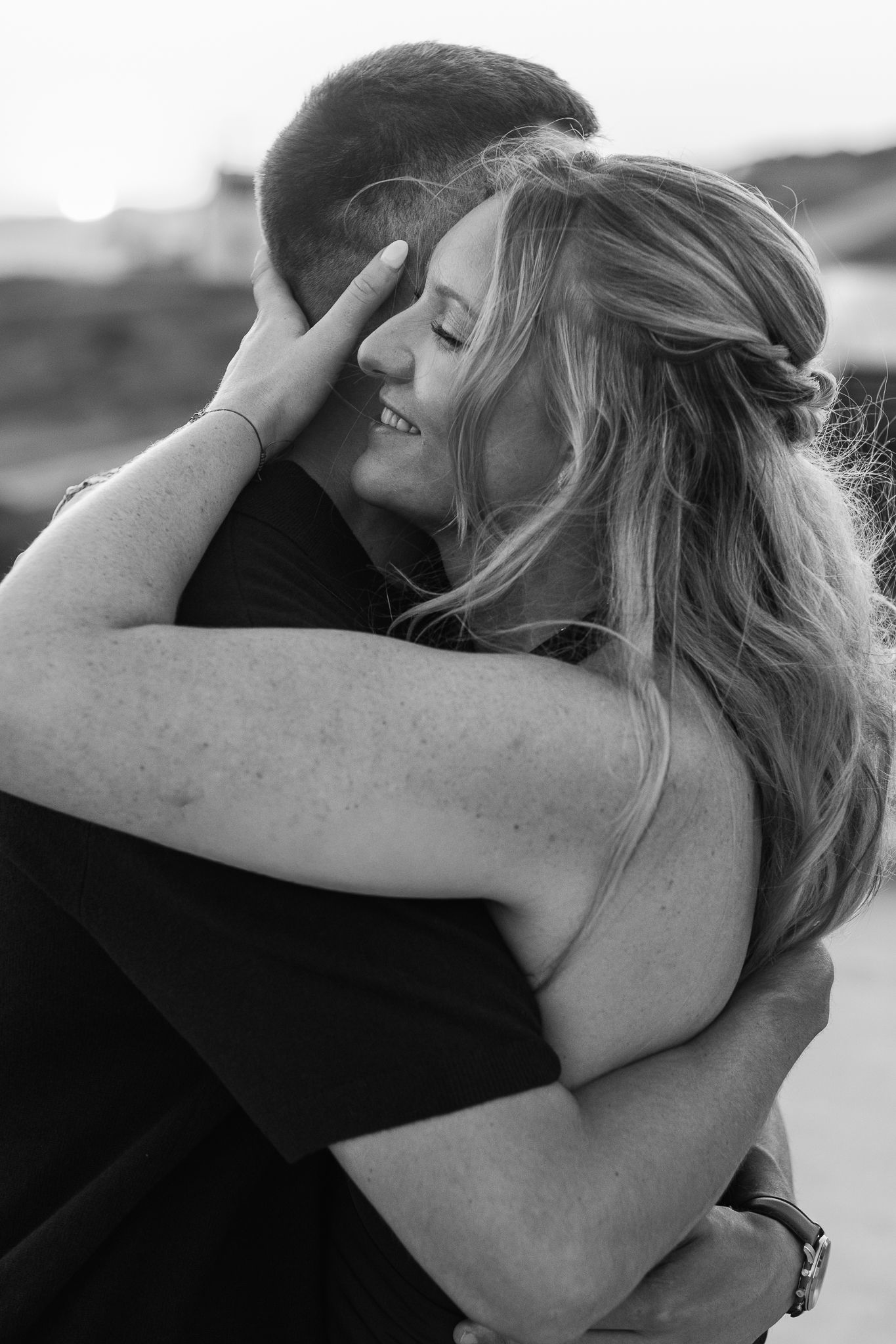 Black and white photo of a smiling woman with long hair embracing a man outdoors in a romantic moment.