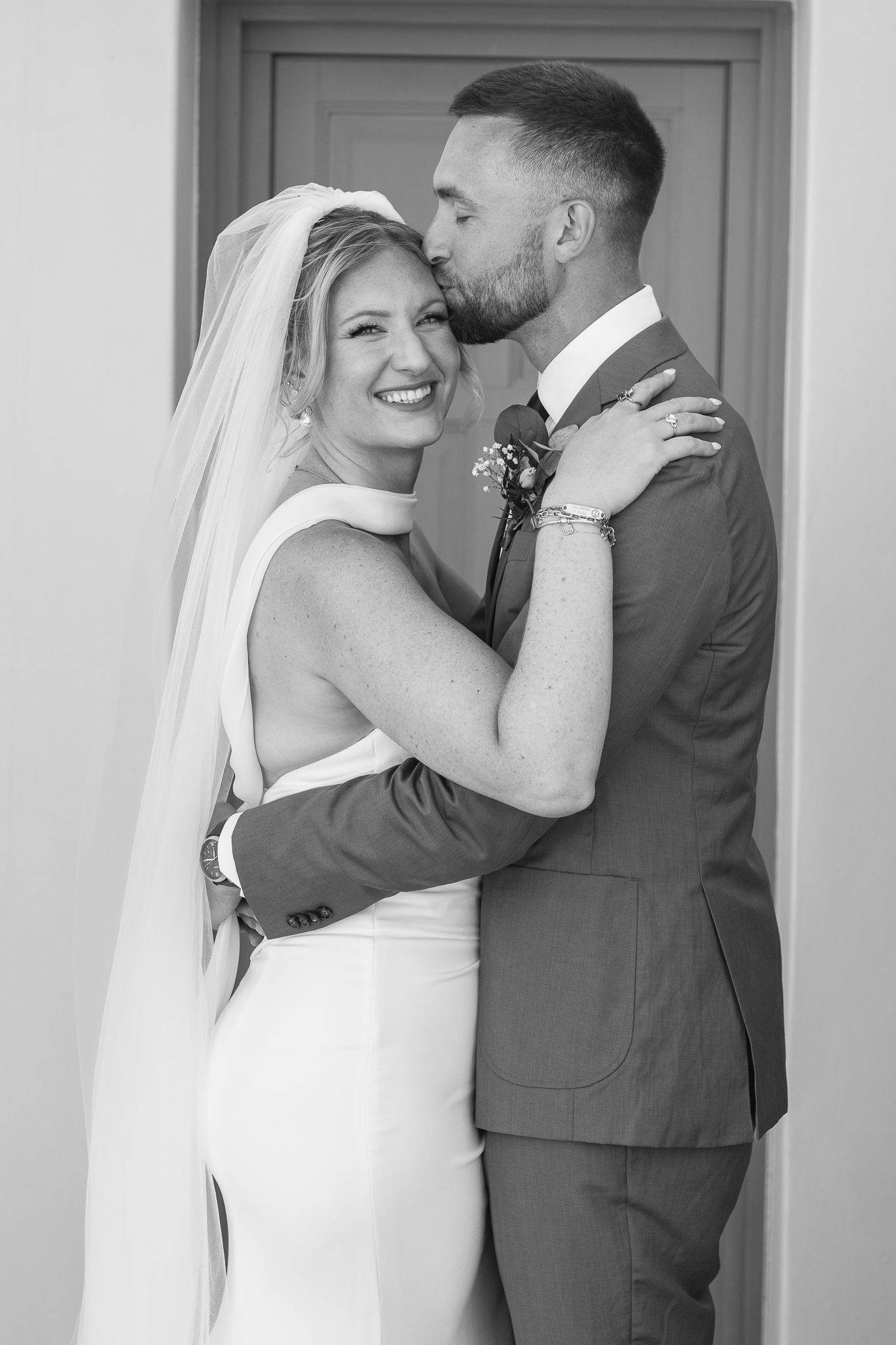 Black and white wedding photo of smiling bride in veil and groom in suit sharing a tender kiss outdoors.