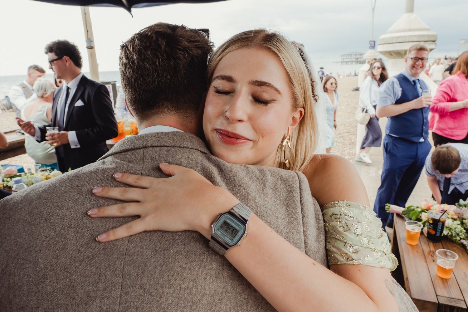 A couple embraces at an outdoor event with guests and floral decorations visible in the background.