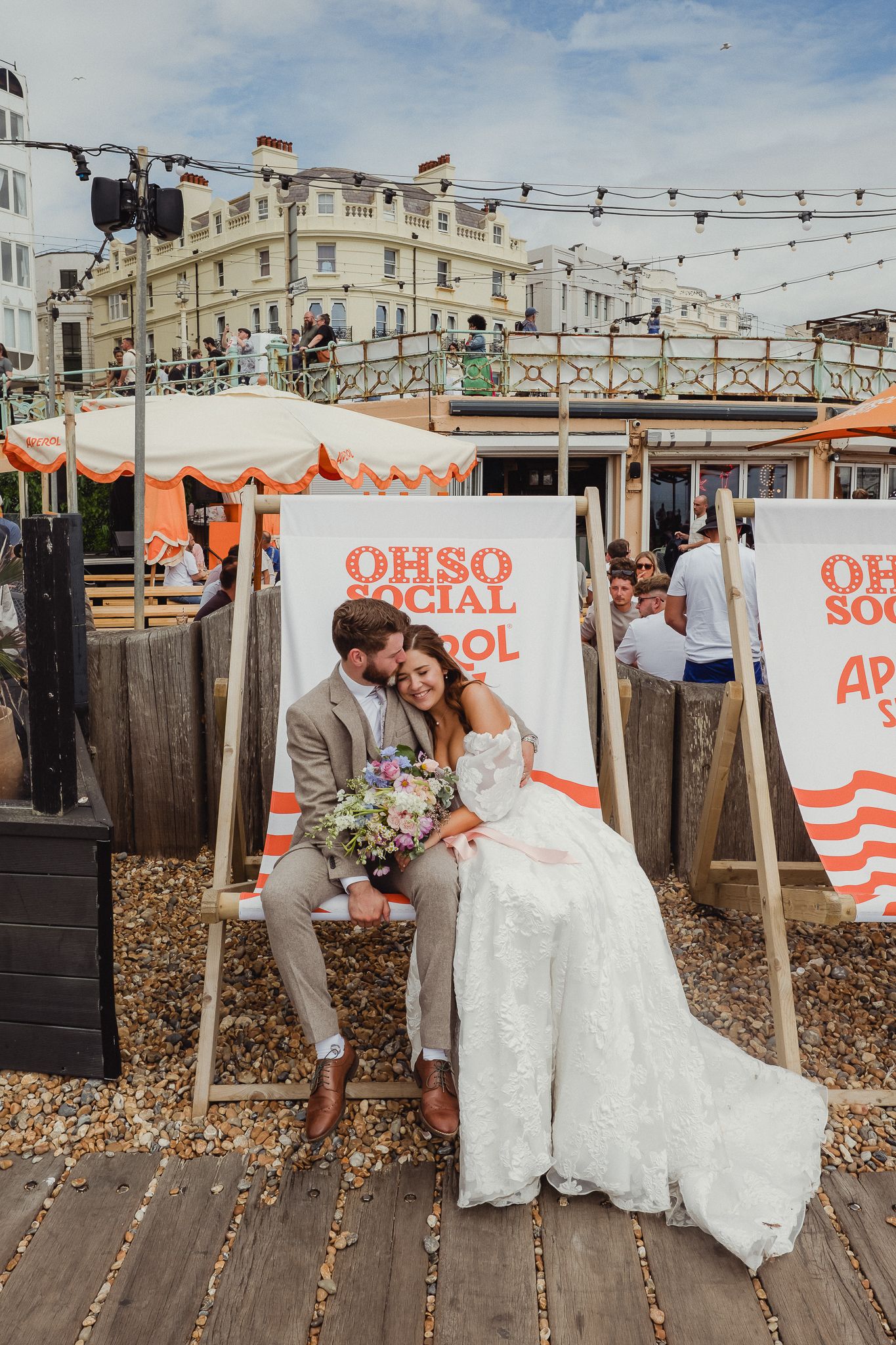 A couple in wedding attire sits on a wooden boardwalk with vintage beach decor and string lights overhead in Brighton.