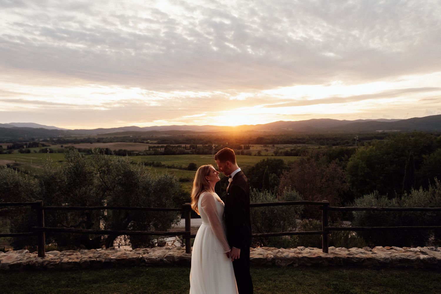 Bride in a white gown and groom in a dark suit share an intimate moment on a stone terrace overlooking rolling hills at golden sunset.