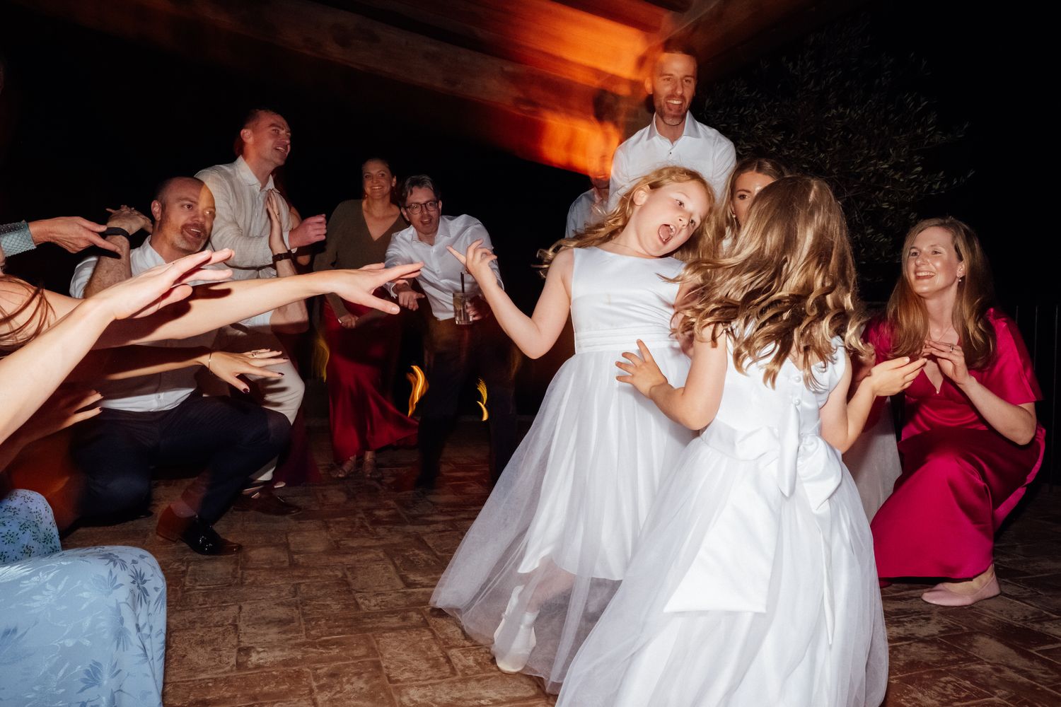 Two flower girls in white dresses dance on the wedding dance floor while smiling guests clap and cheer around them.