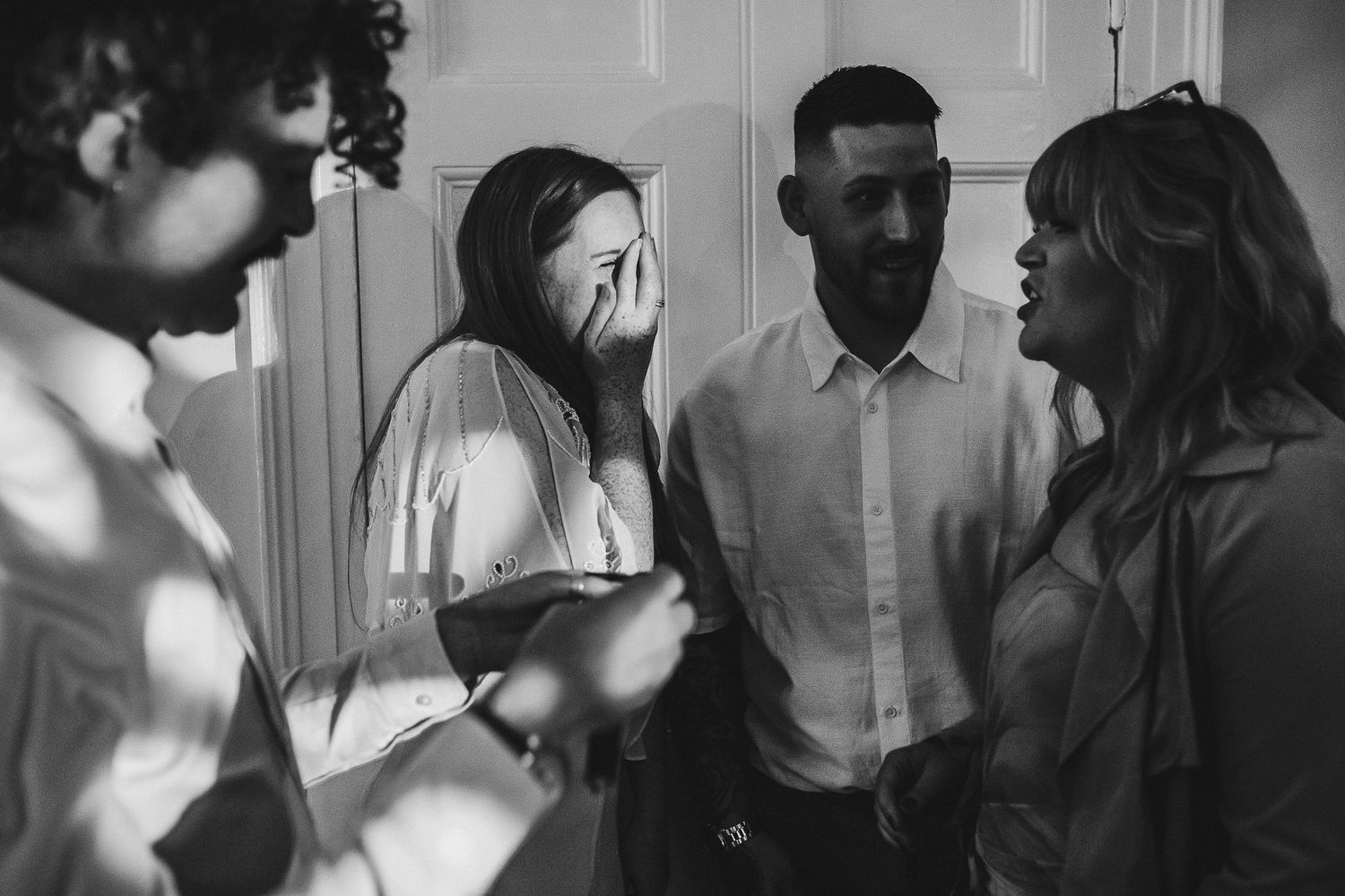 Black and white photo of four people in an emotional moment, with one woman covering her face while others look on in a bright room.