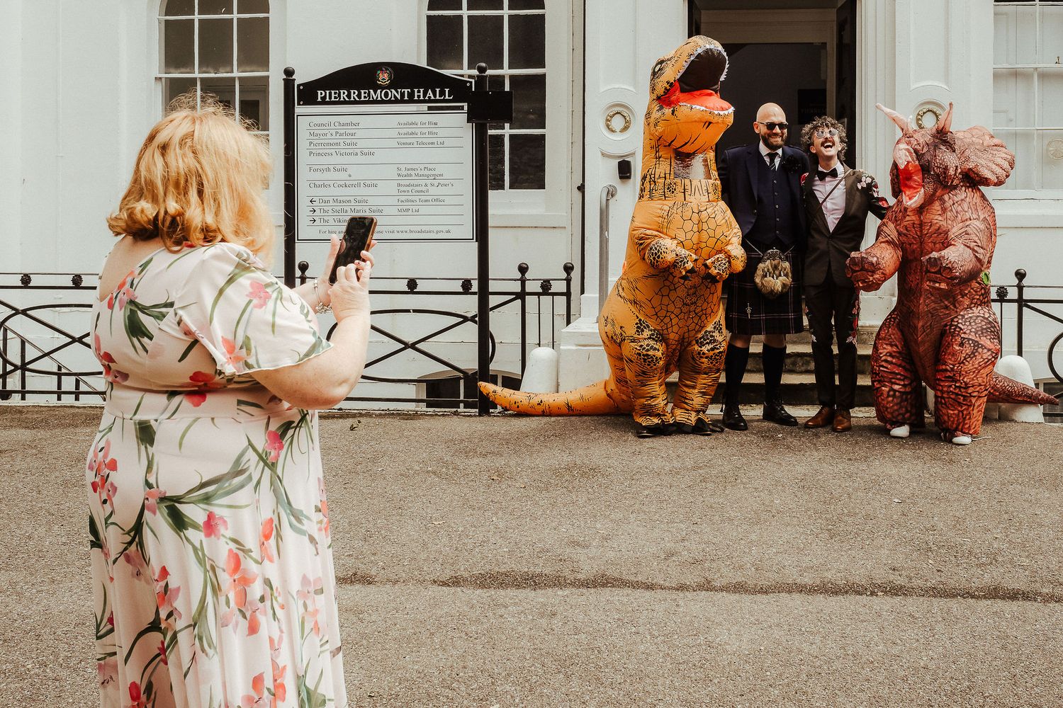 Woman in a floral dress photographs two people in inflatable dinosaur costumes posing with a smiling couple outside a stately building.