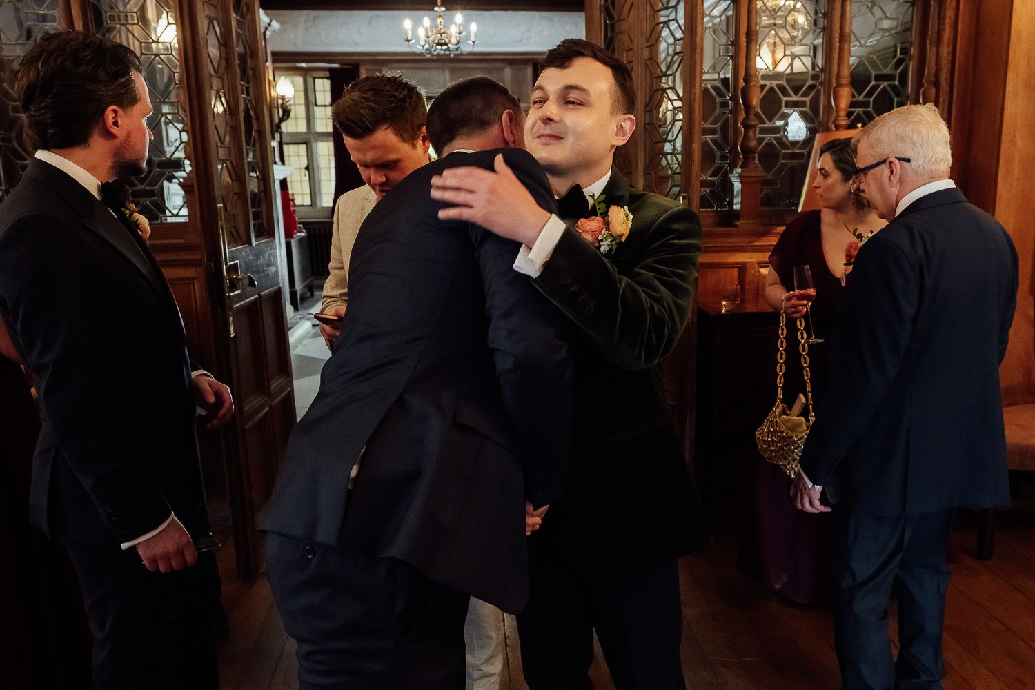 Men in formal suits hugging and socializing at an elegant wedding reception in a wood-paneled room.