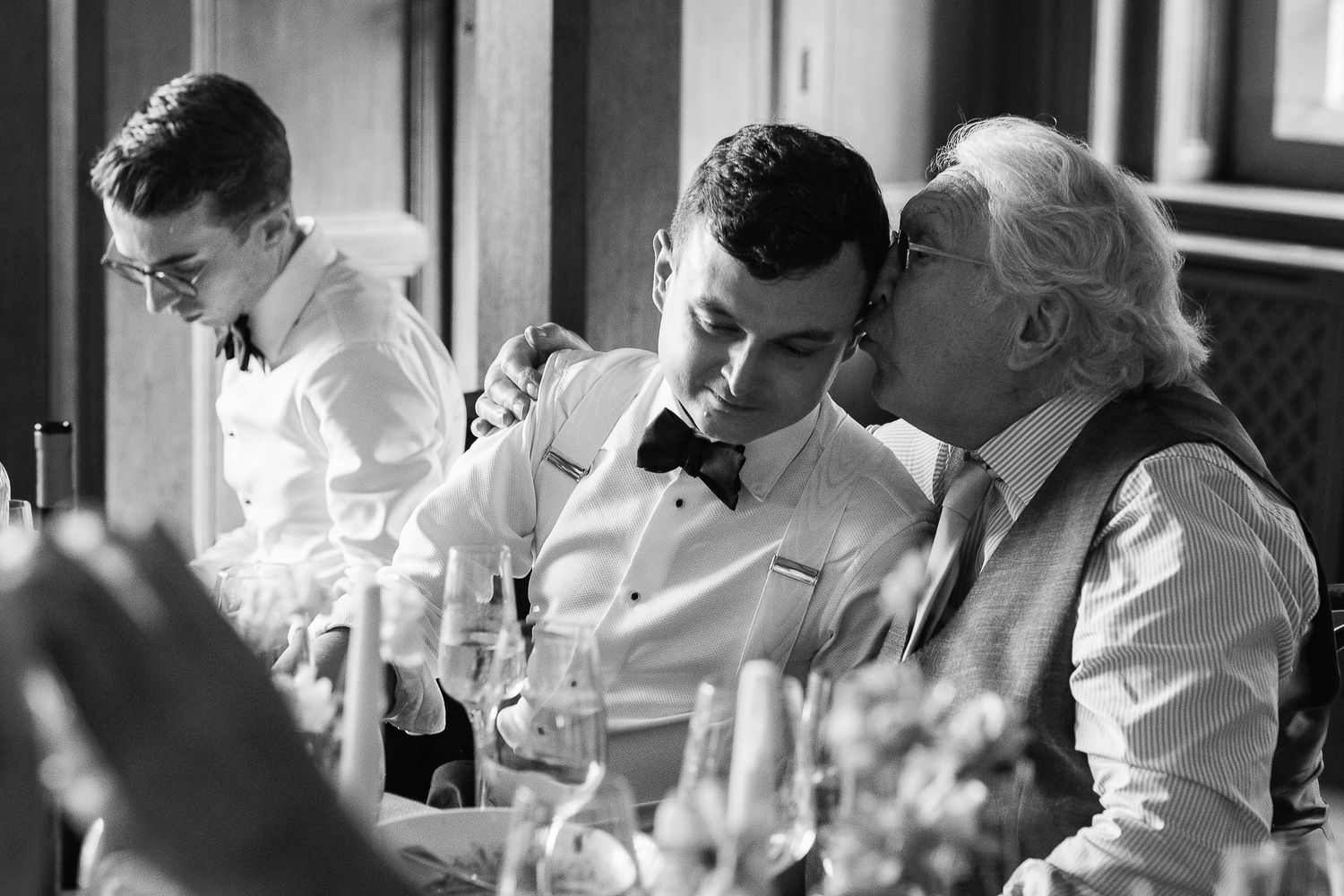 Black and white wedding photo of groom receiving a kiss on cheek from elder guest at elegant reception dinner.