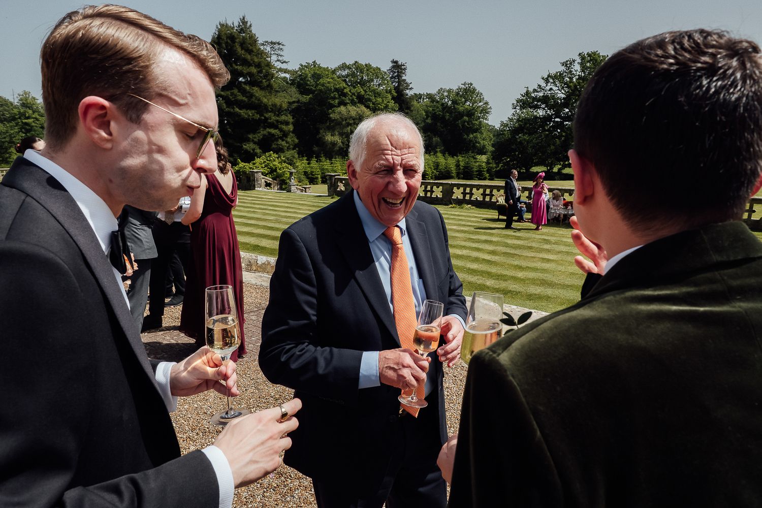 Elegantly dressed guests socializing outdoors at a garden wedding reception, holding champagne glasses on a sunny day.
