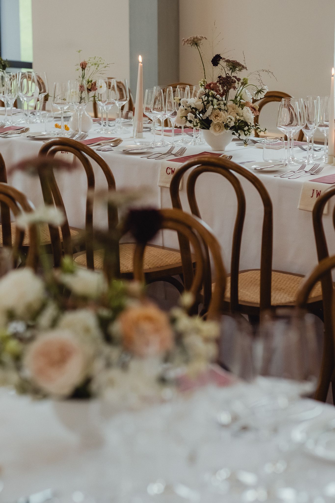 Elegant wedding reception tables set with white linens, floral centerpieces and dark wood chairs in a bright dining room.