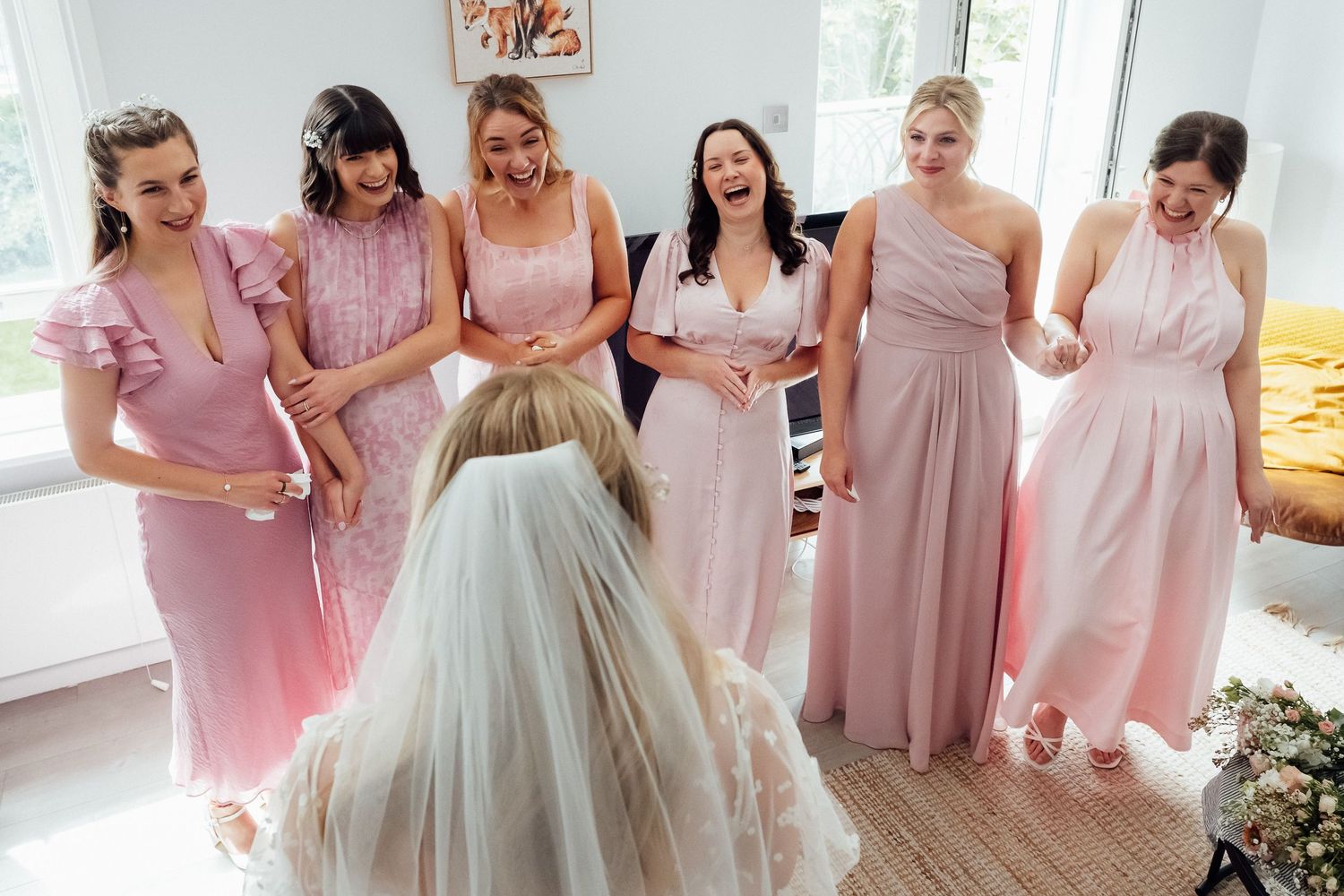 Six bridesmaids in blush pink dresses smile and laugh during a joyful first look with the bride, who wears a veil and faces away.