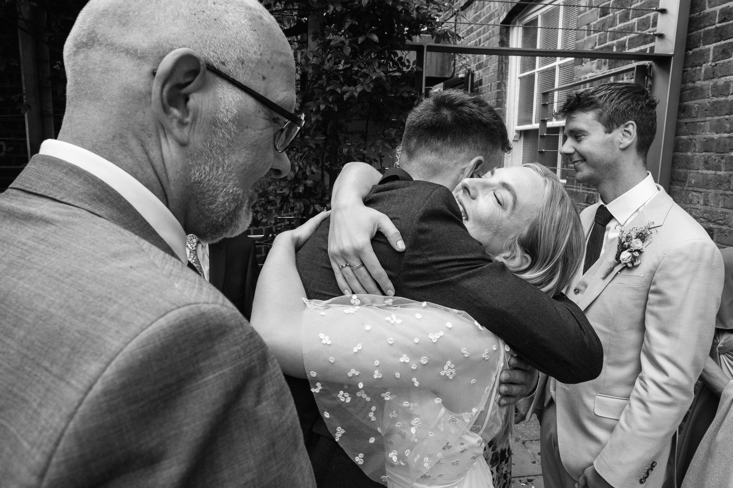 Black and white wedding photo of a bride in a lace dress hugging a man in a suit while an older man and a groomsman look on.