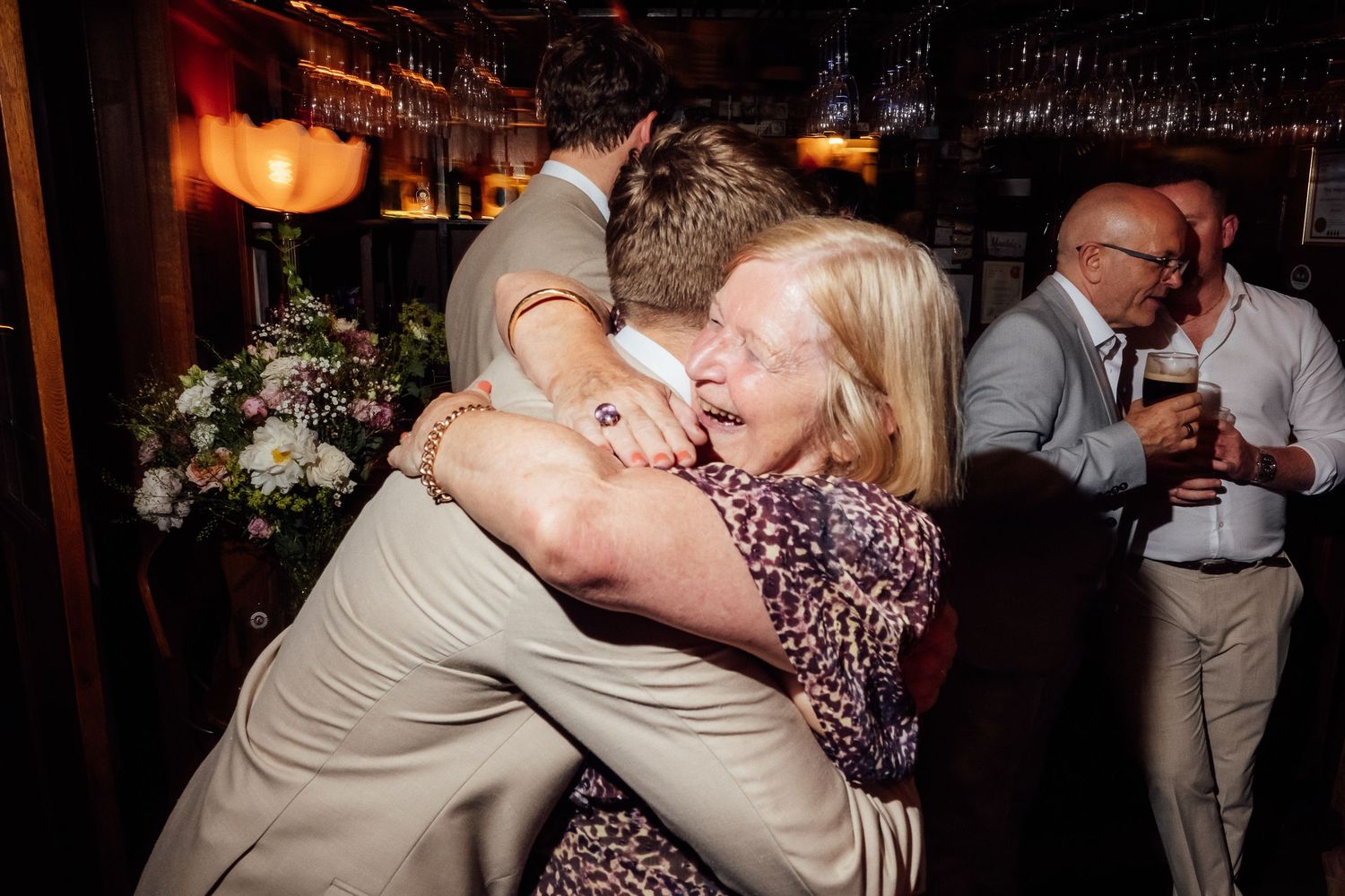 Smiling older woman with blonde hair hugs a man in a tan suit at a lively indoor celebration while guests chat near a warmly lit bar.