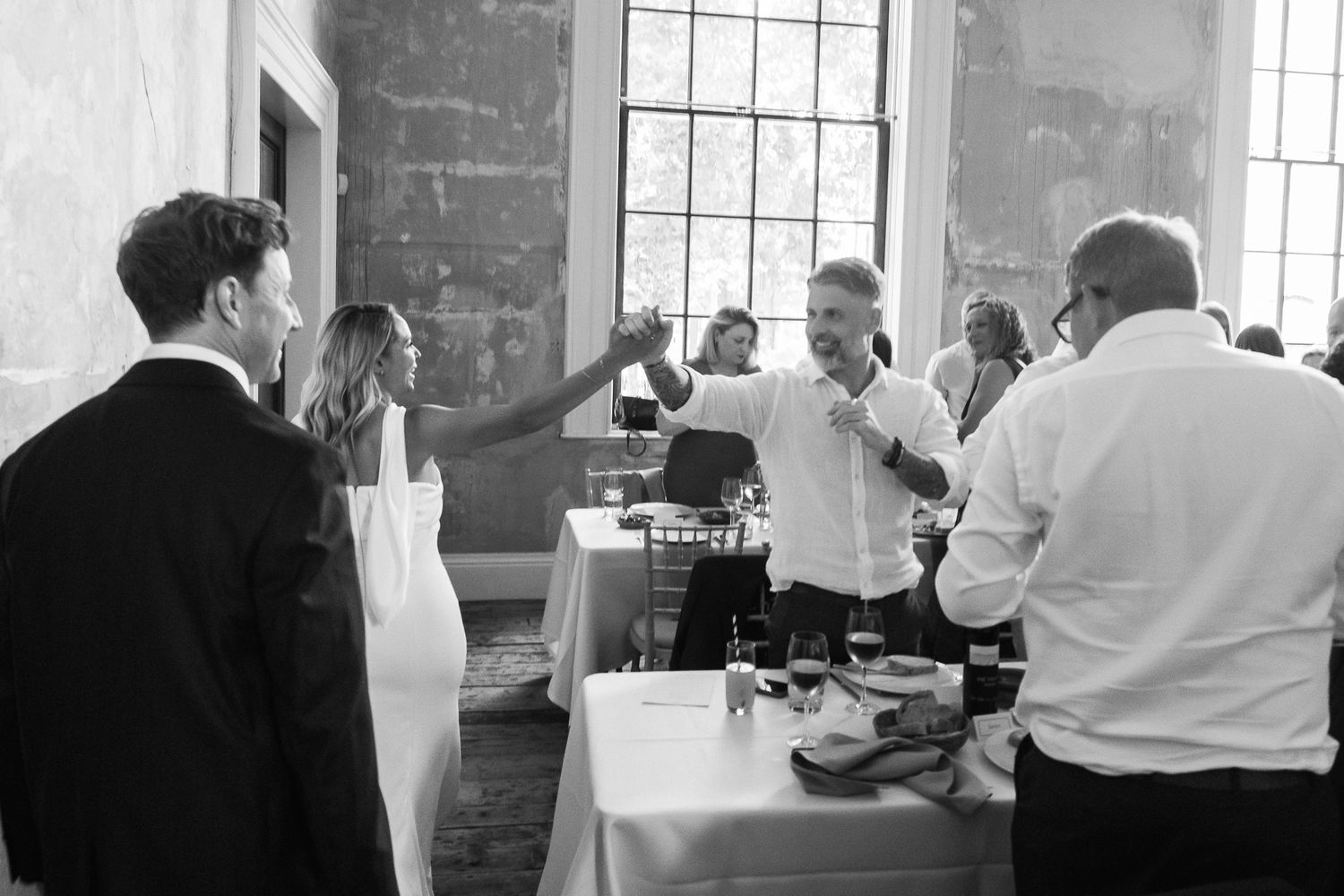 Wedding guests raise their glasses in a toast at a reception with white-clothed tables inside a bright venue with tall windows.