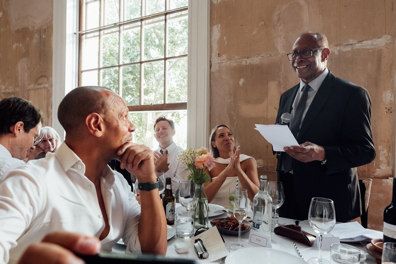 Man in a dark suit holds a paper and gives a speech at a wedding reception table while seated guests listen and smile.