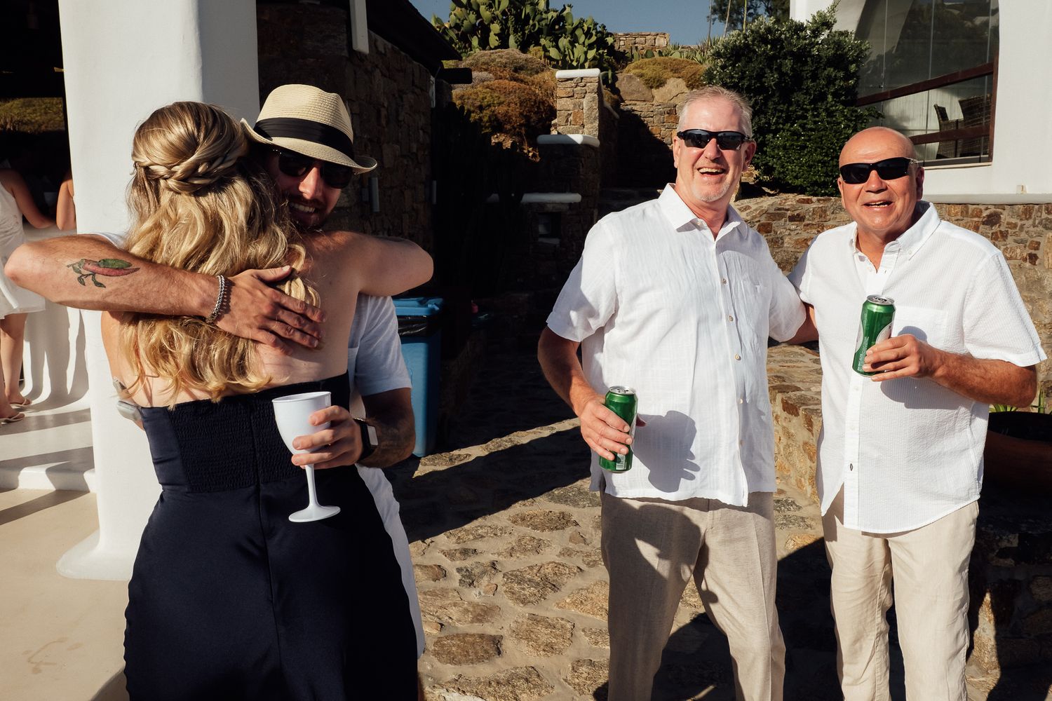 Guests hugging and socializing outdoors at a sunny wedding reception, two men smiling and holding green beer bottles.