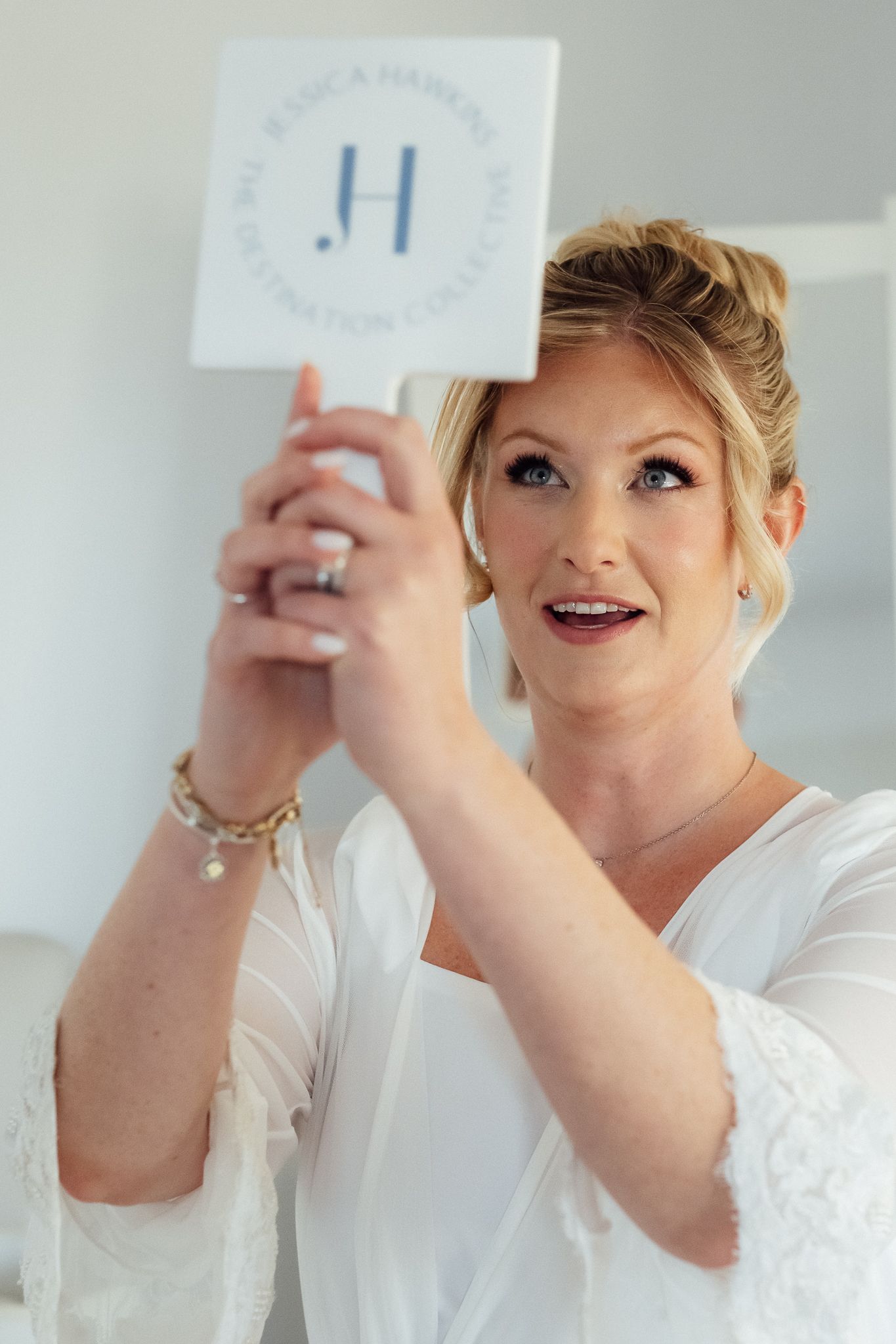 Blonde woman in white robe holding a JH signature collection card, wearing jewelry, with elegant updo hairstyle.