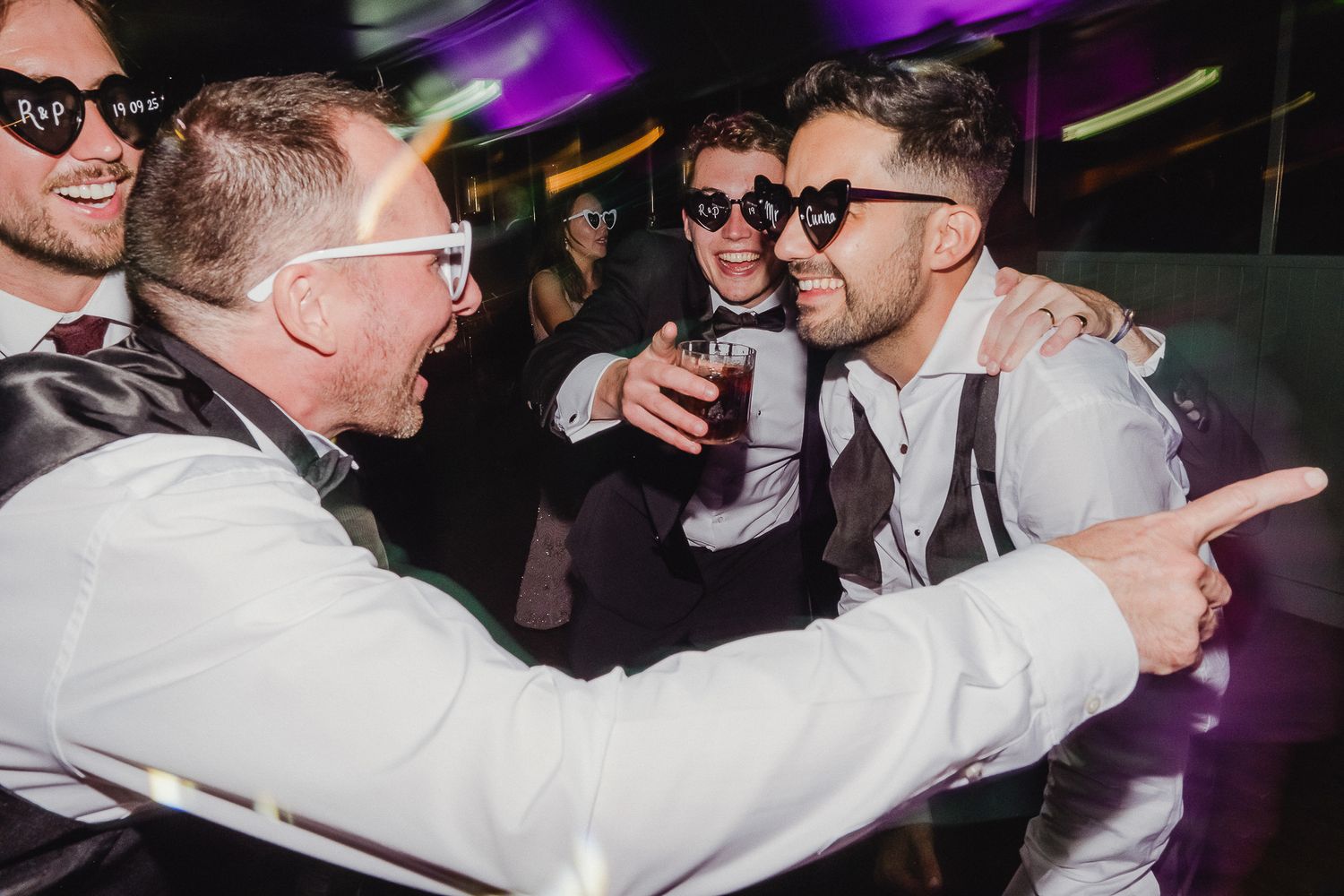 Party guests in sunglasses and formal attire celebrate on a dance floor under purple lighting.