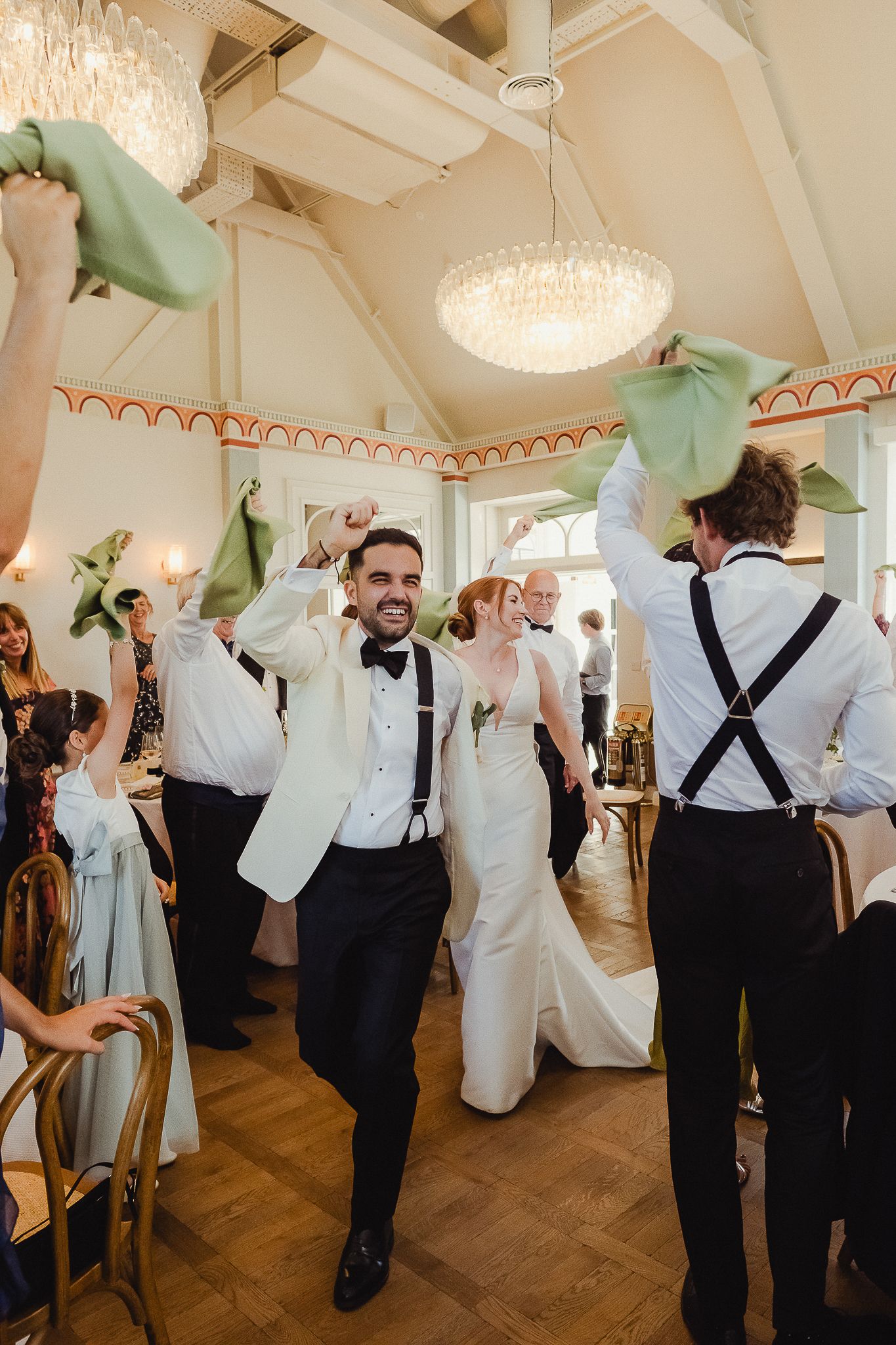 Wedding reception celebration in an elegant ballroom with chandelier and guests waving mint green napkins on parquet floors.