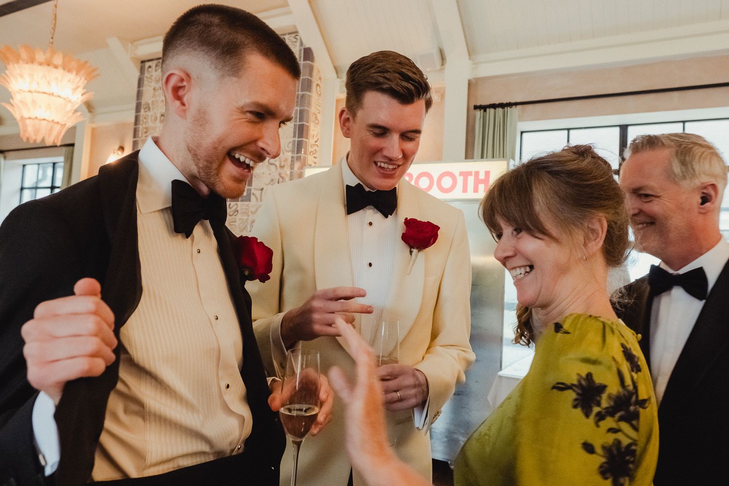 A group of formally dressed guests share a joyful moment at an elegant wedding reception with a chandelier in the background.