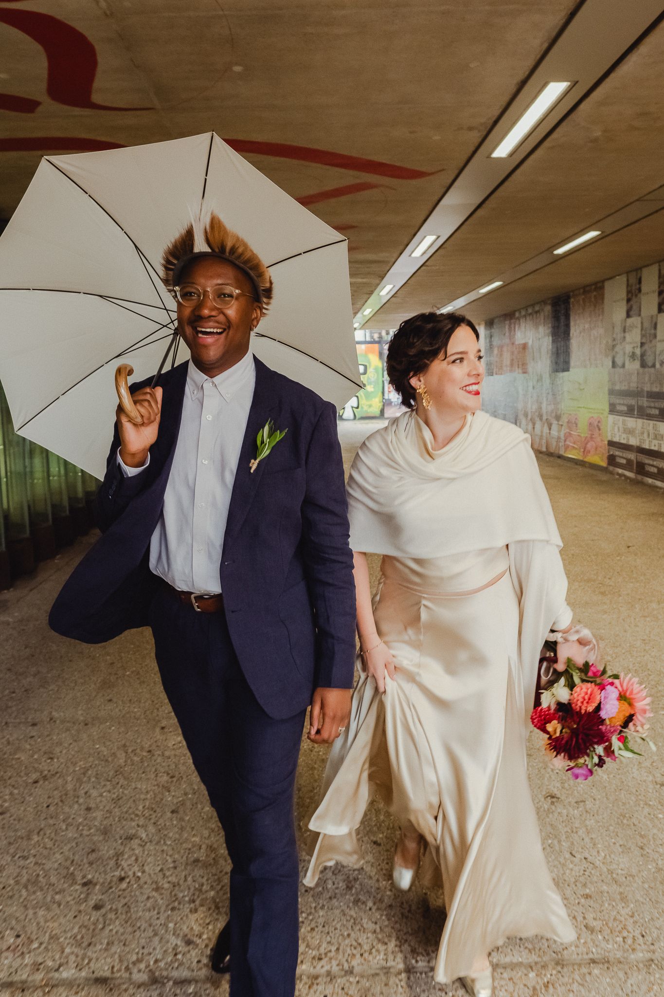 A couple shares a joyful moment under a concrete structure while dressed in wedding attire.