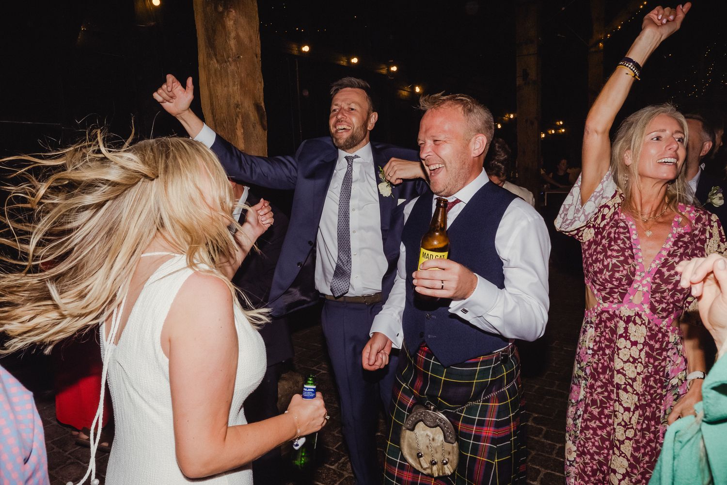 Wedding guests dance energetically at a reception with blonde hair swinging and a man in traditional Scottish kilt.