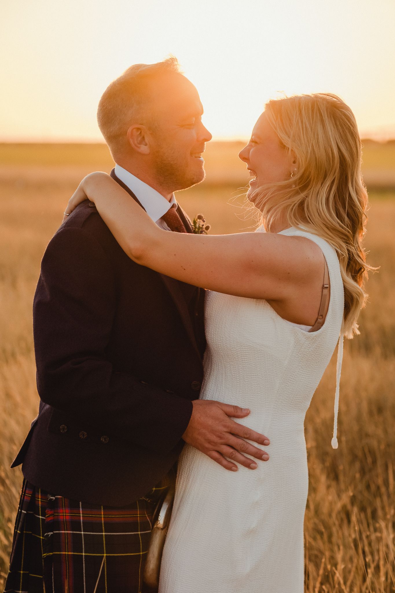 A couple embraces in a golden wheat field at sunset while wearing formal wedding attire.
