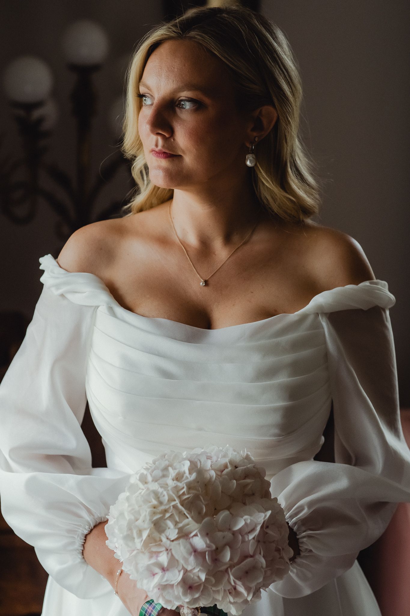 A bride in an elegant white off-shoulder wedding dress with puff sleeves holds a white peony bouquet.