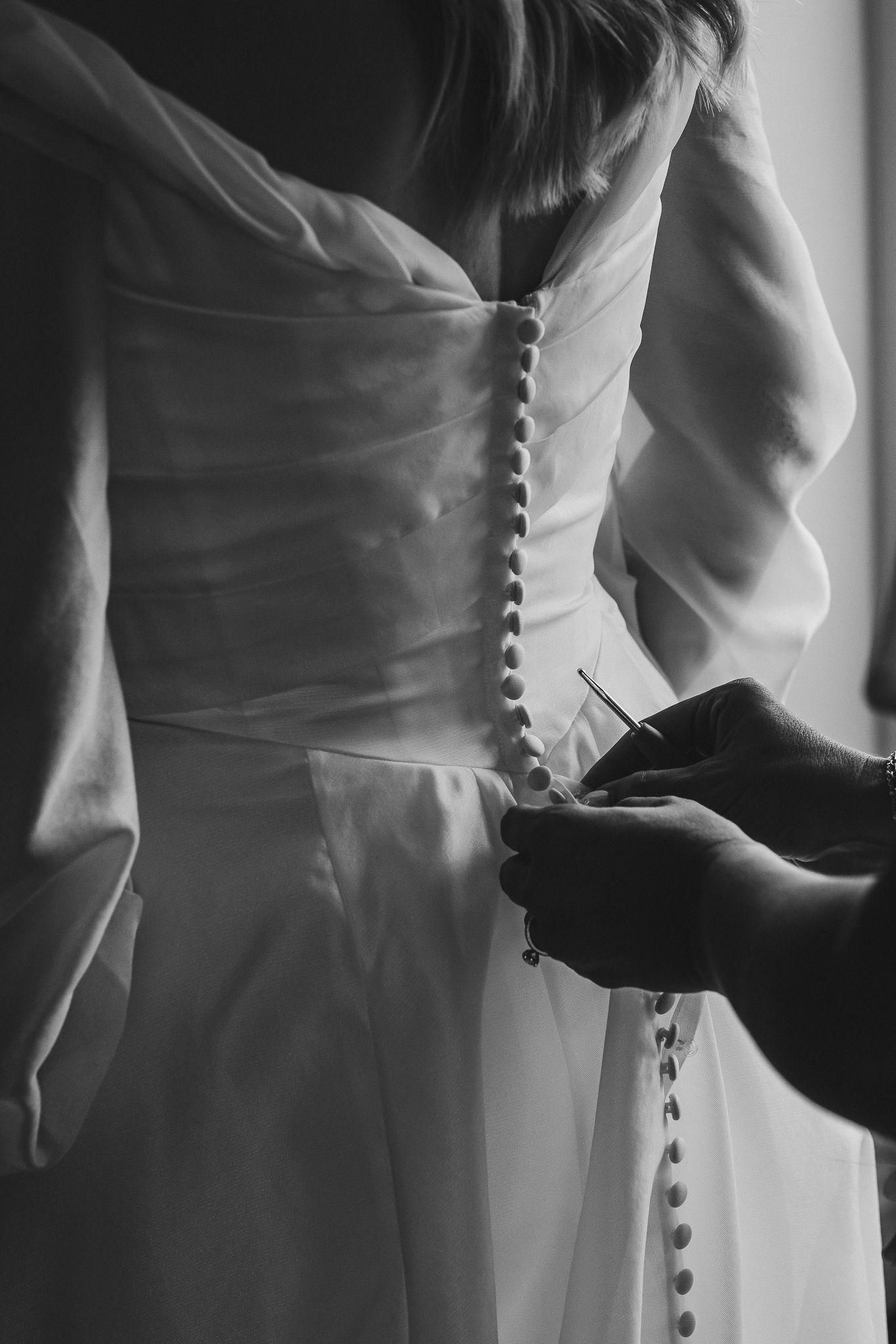 Black and white detail shots of buttons being fastened on the back of an elegant white wedding dress.