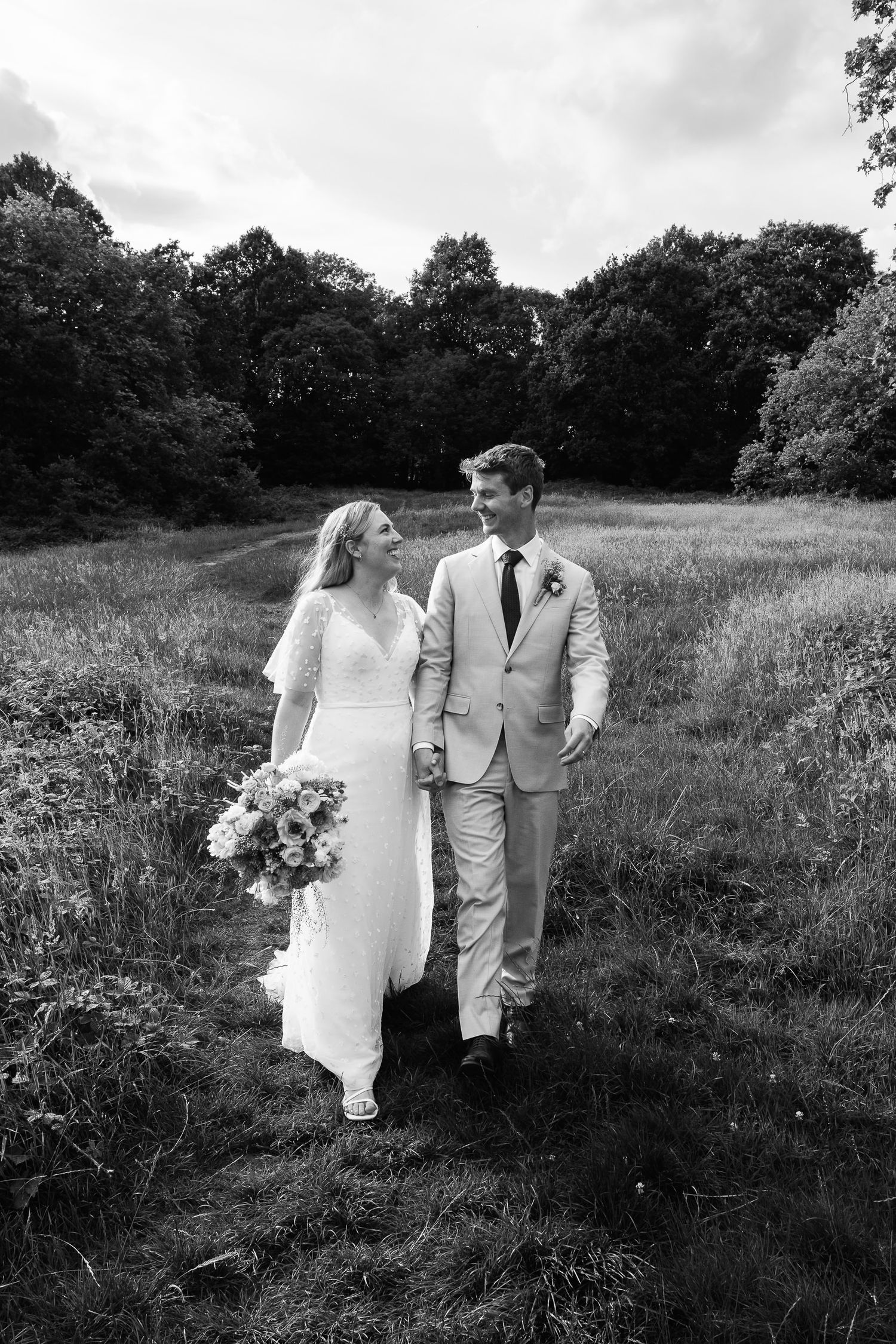 Bride in a lace wedding dress holding a bouquet walks hand in hand with a groom in a light suit through a grassy meadow.