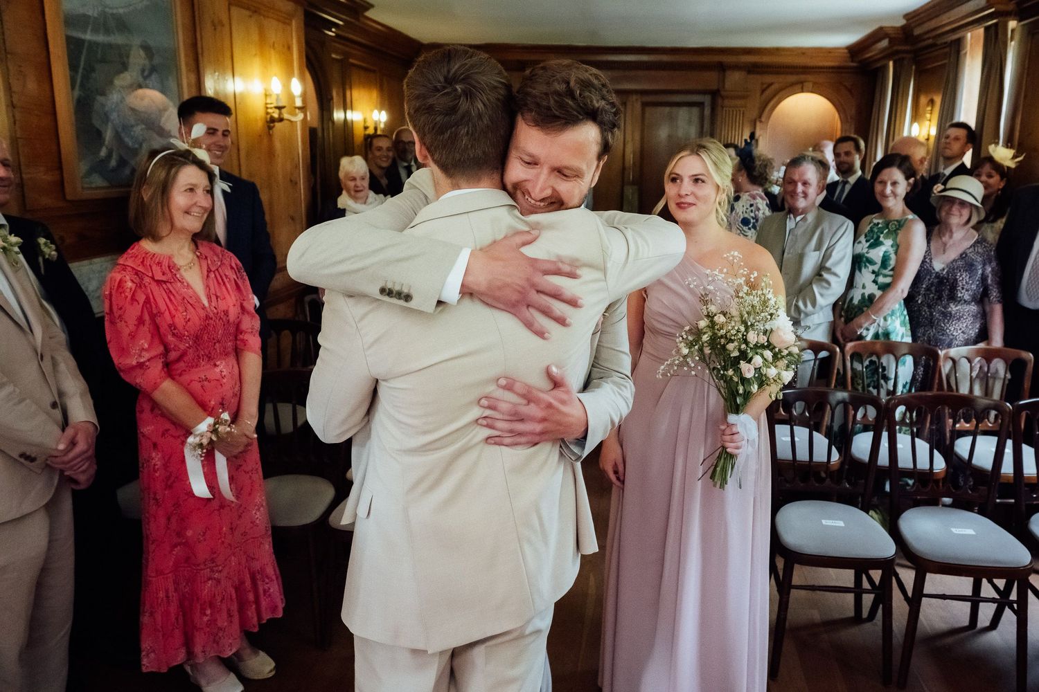 Two men in light suits share an emotional hug during a wedding ceremony while guests look on and a woman holds a bouquet.