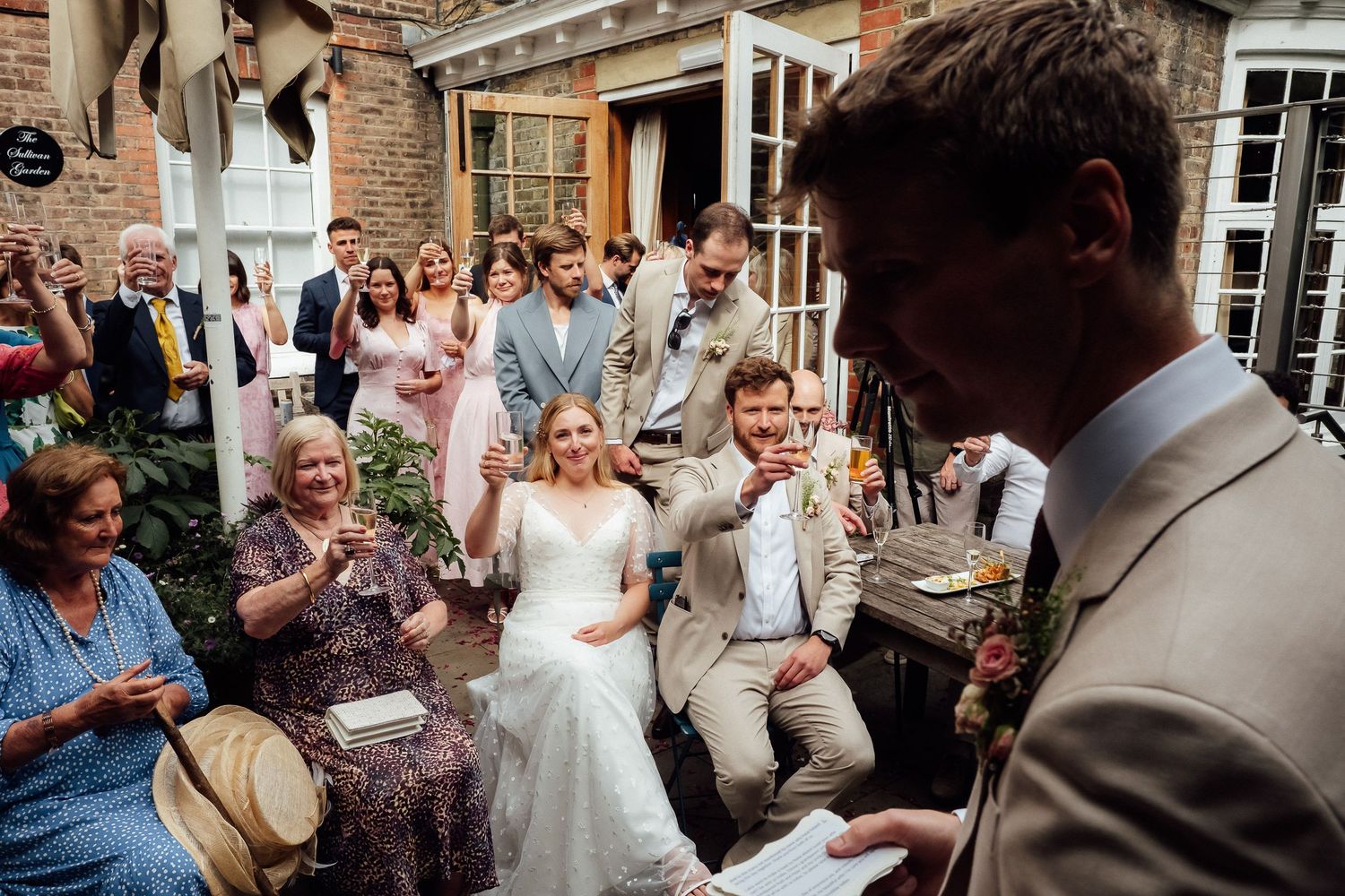 Bride in a white lace gown smiles joyfully among seated wedding guests as a groom in a tan suit reads vows at an outdoor brick venue.