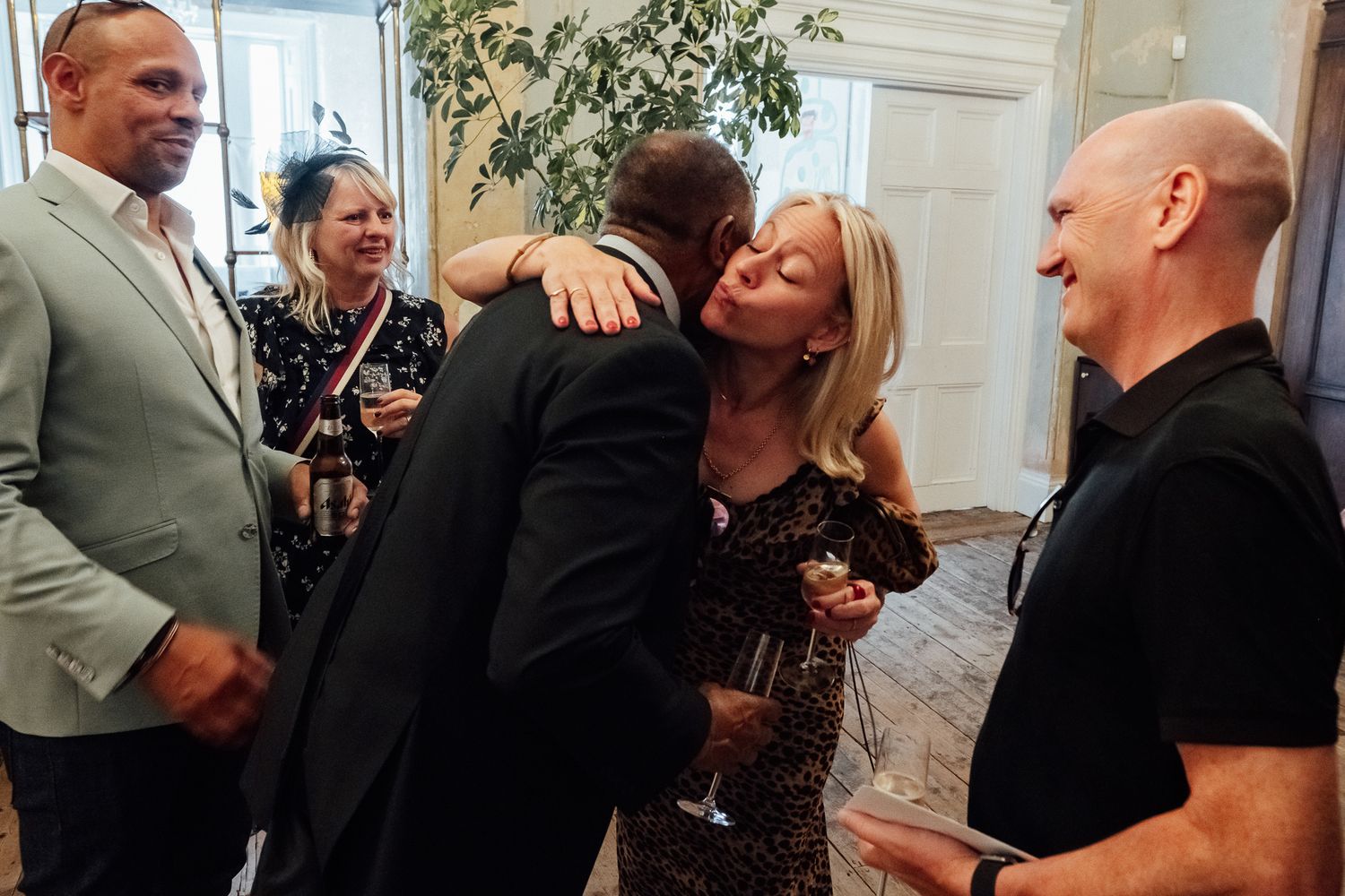 Guests embrace and chat at an indoor gathering while holding drinks, with smiling attendees looking on in a bright, elegant room.