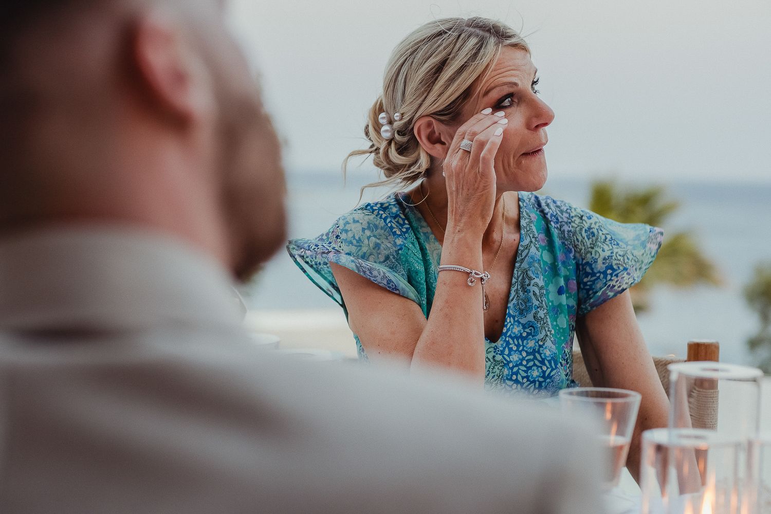 Person wearing a turquoise sequined dress and updo hairstyle sits at an outdoor table overlooking an ocean view.