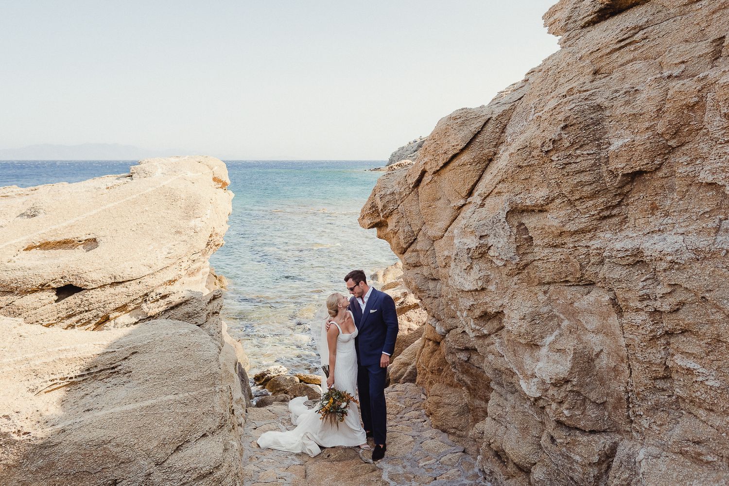 A couple embraces on a rocky coastal cliff overlooking pristine turquoise waters.