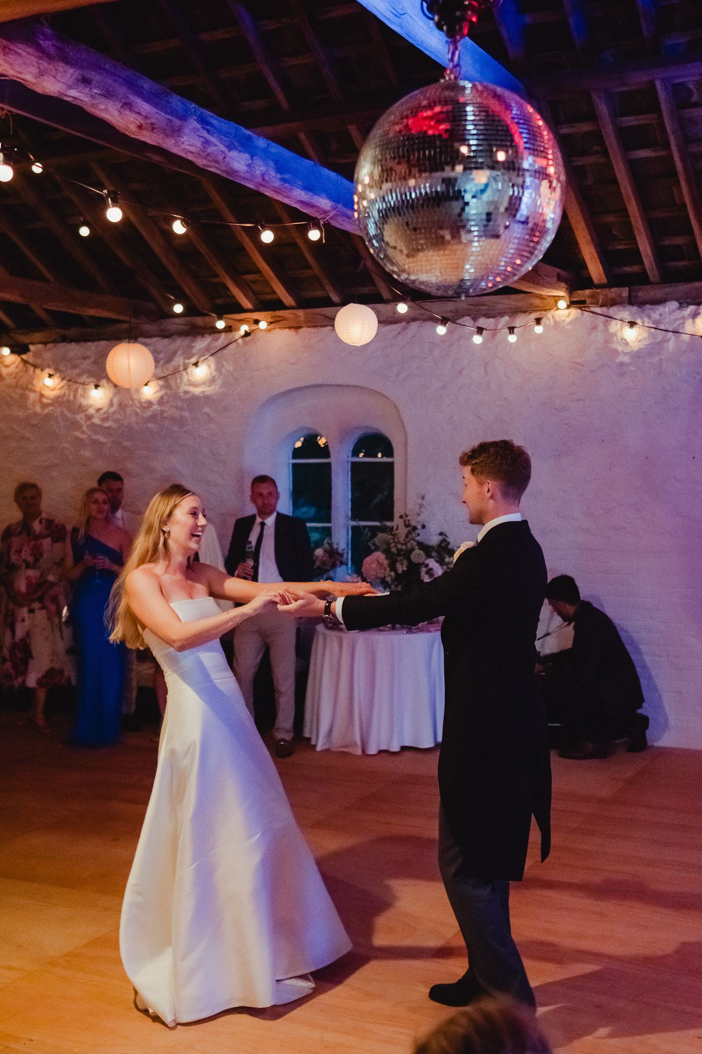 A couple dances on a wooden floor under a disco ball with white walls and twinkling lights in the background.