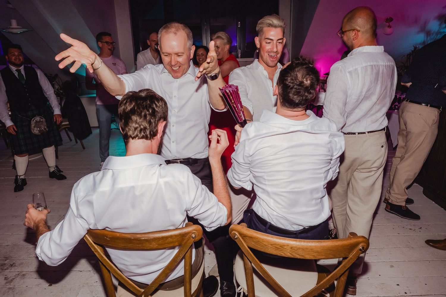 Party guests in white shirts dance and interact at a celebration with wooden chairs and colorful lighting in the background.