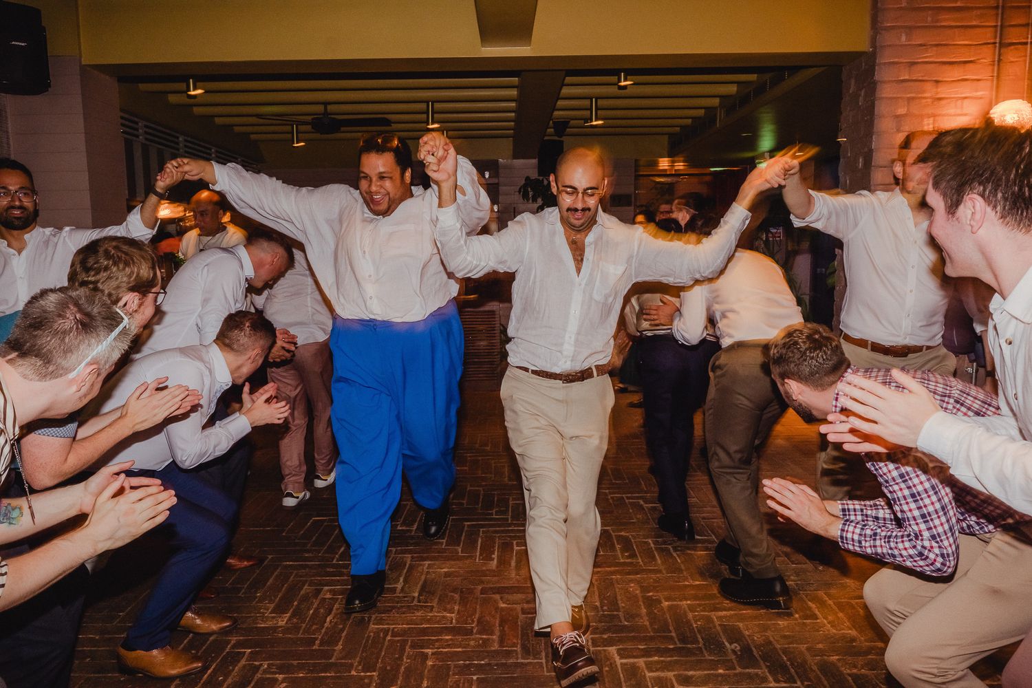 A line of wedding guests on a brick floor performing a synchronized dance move during a reception celebration.