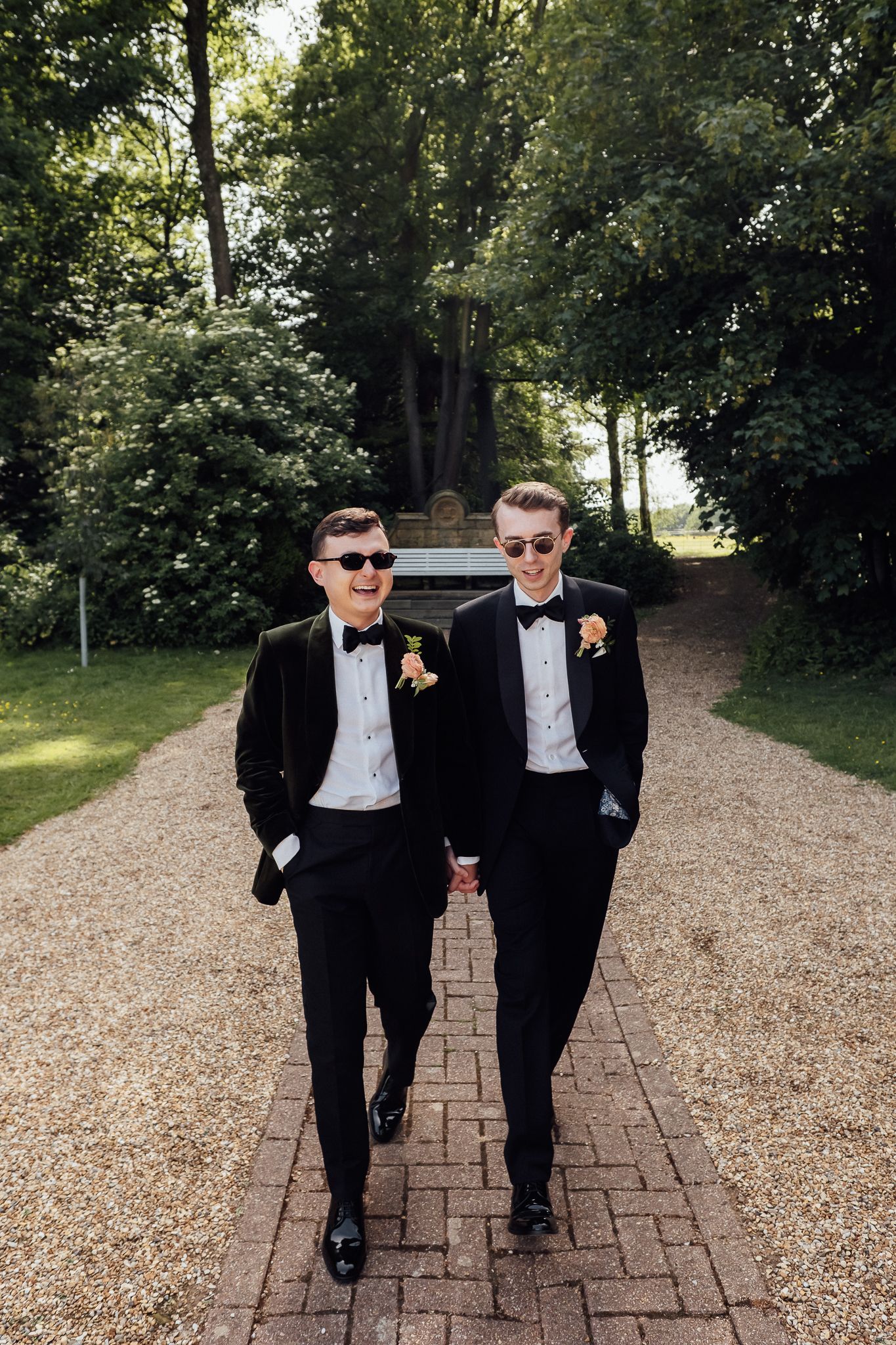 Two grooms in black tuxedos and sunglasses walk hand-in-hand down a garden path on their wedding day.