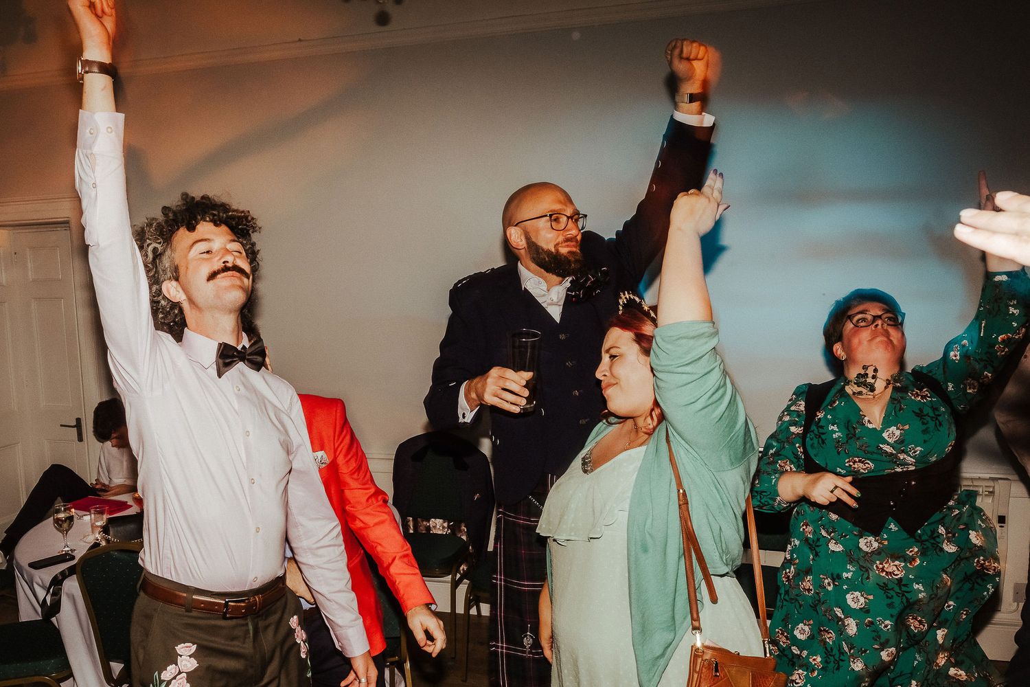 Wedding guests in festive attire dance joyfully with raised fists and arms at a lively indoor reception party.