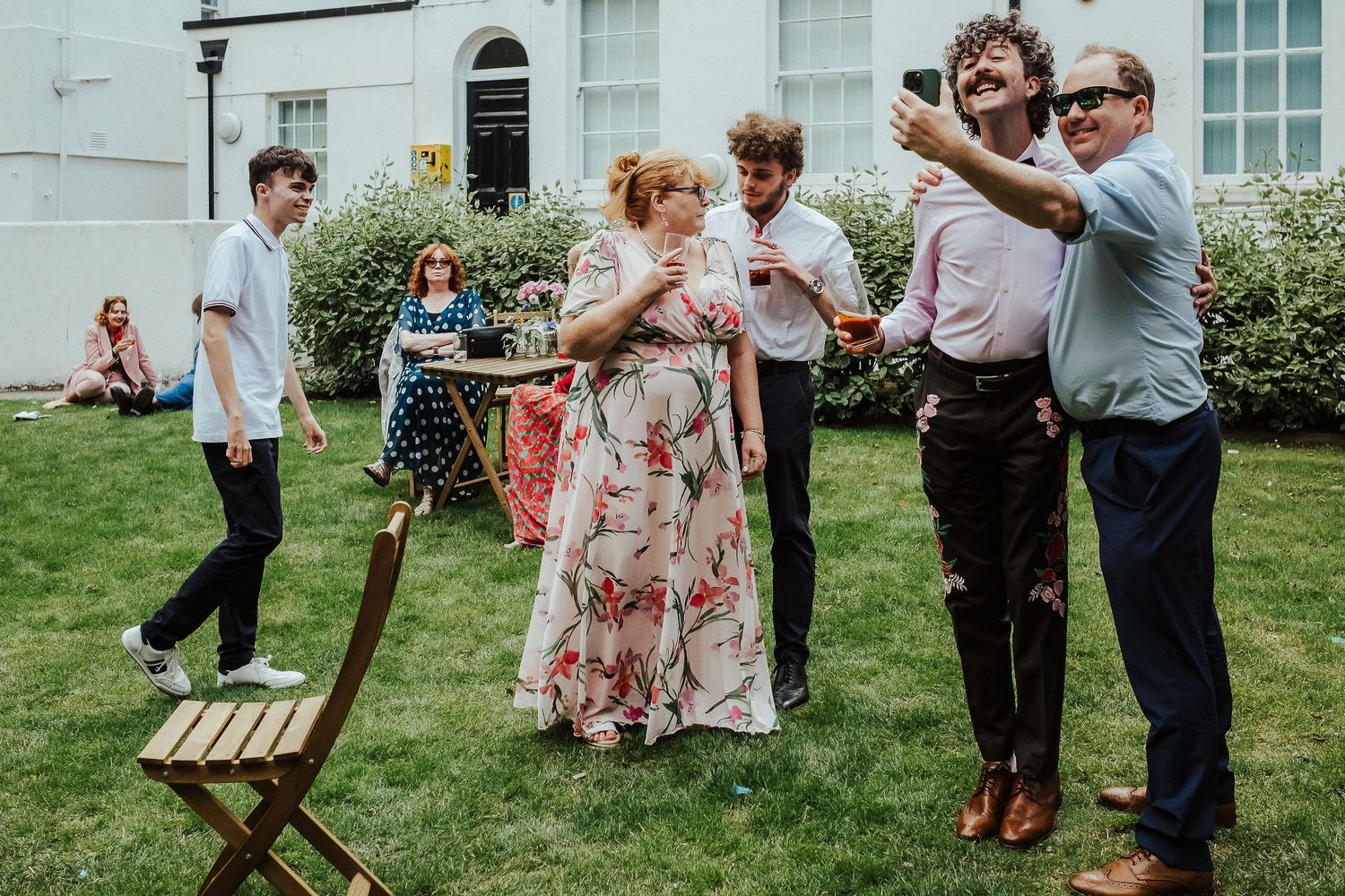 Guests in summer clothes laugh and take a group selfie with drinks on a sunny garden lawn beside a wooden folding chair.