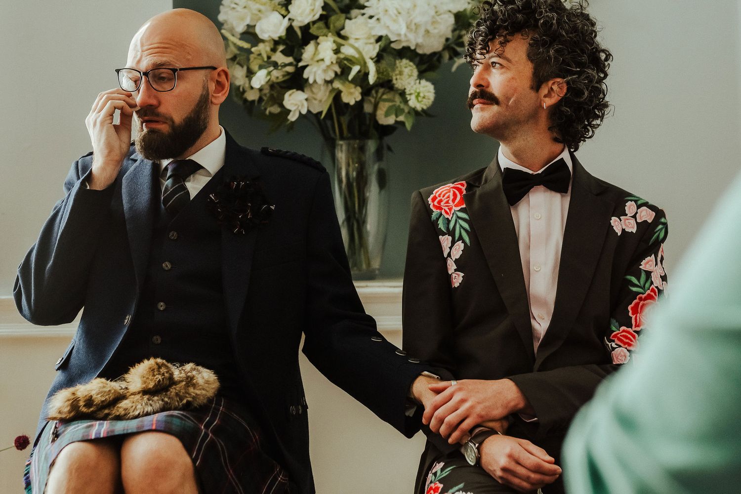 Emotional groom in glasses wipes a tear while holding hands with his partner in a floral blazer and bow tie at a wedding ceremony.