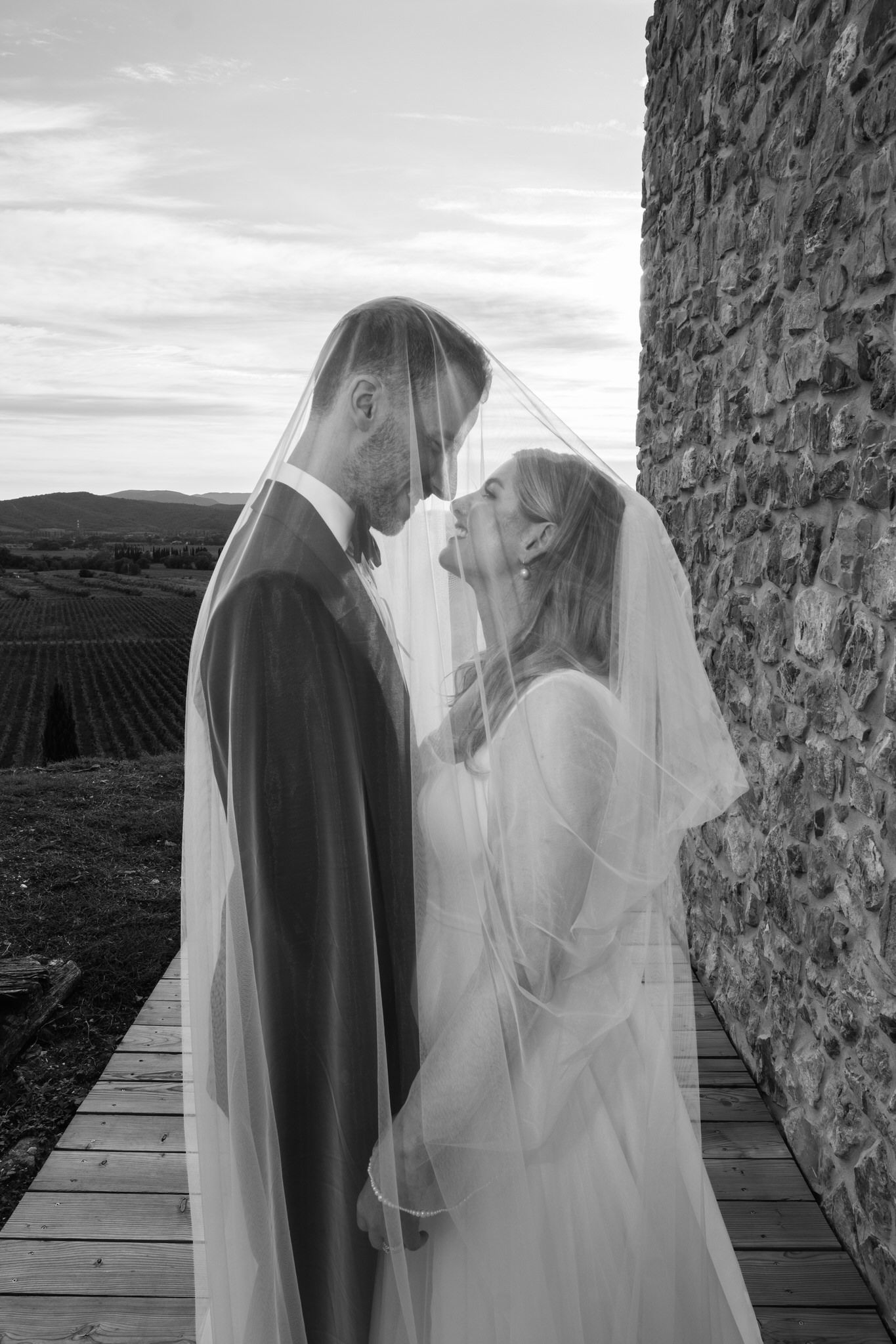 Black and white wedding portrait of a couple touching foreheads under a veil beside a stone wall and vineyard.