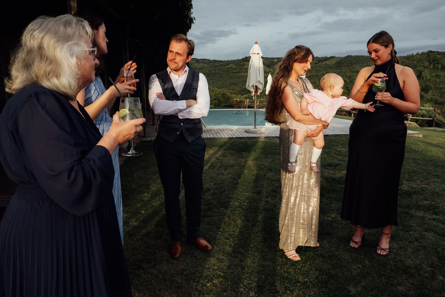 Wedding guests in formal attire hold champagne glasses and chat on a lawn at dusk, with a woman cradling a baby near a pool.