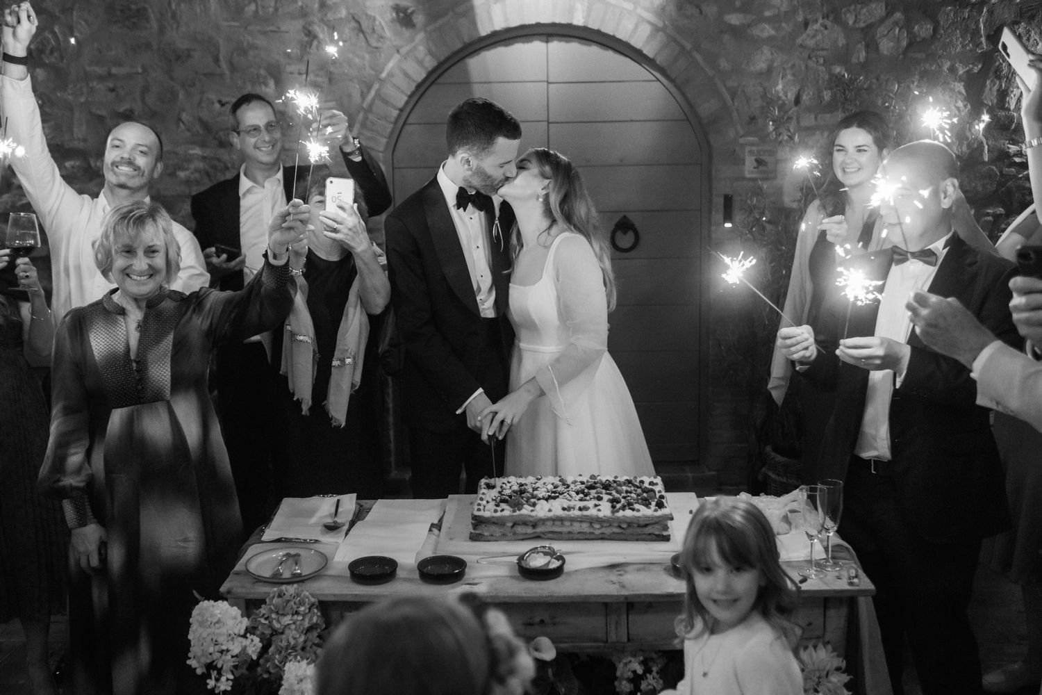 Bride and groom share an intimate moment beside their wedding cake as guests cheer, film, and hold sparklers at a rustic stone venue.