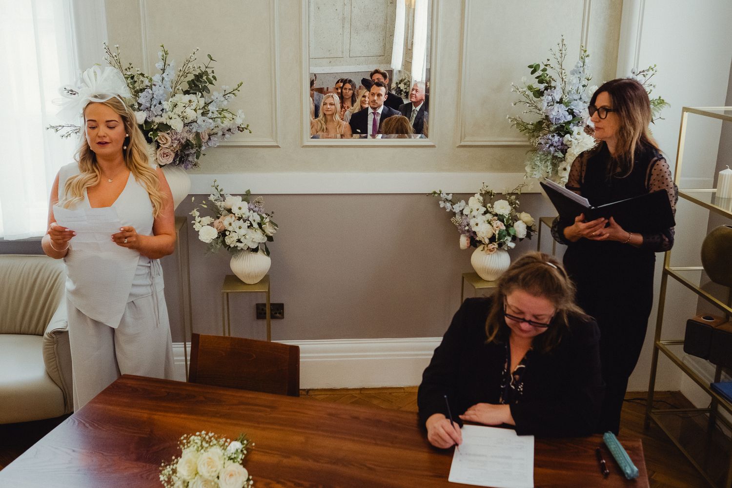 A wedding preparation scene in a room with white floral arrangements and people gathered around a wooden table.